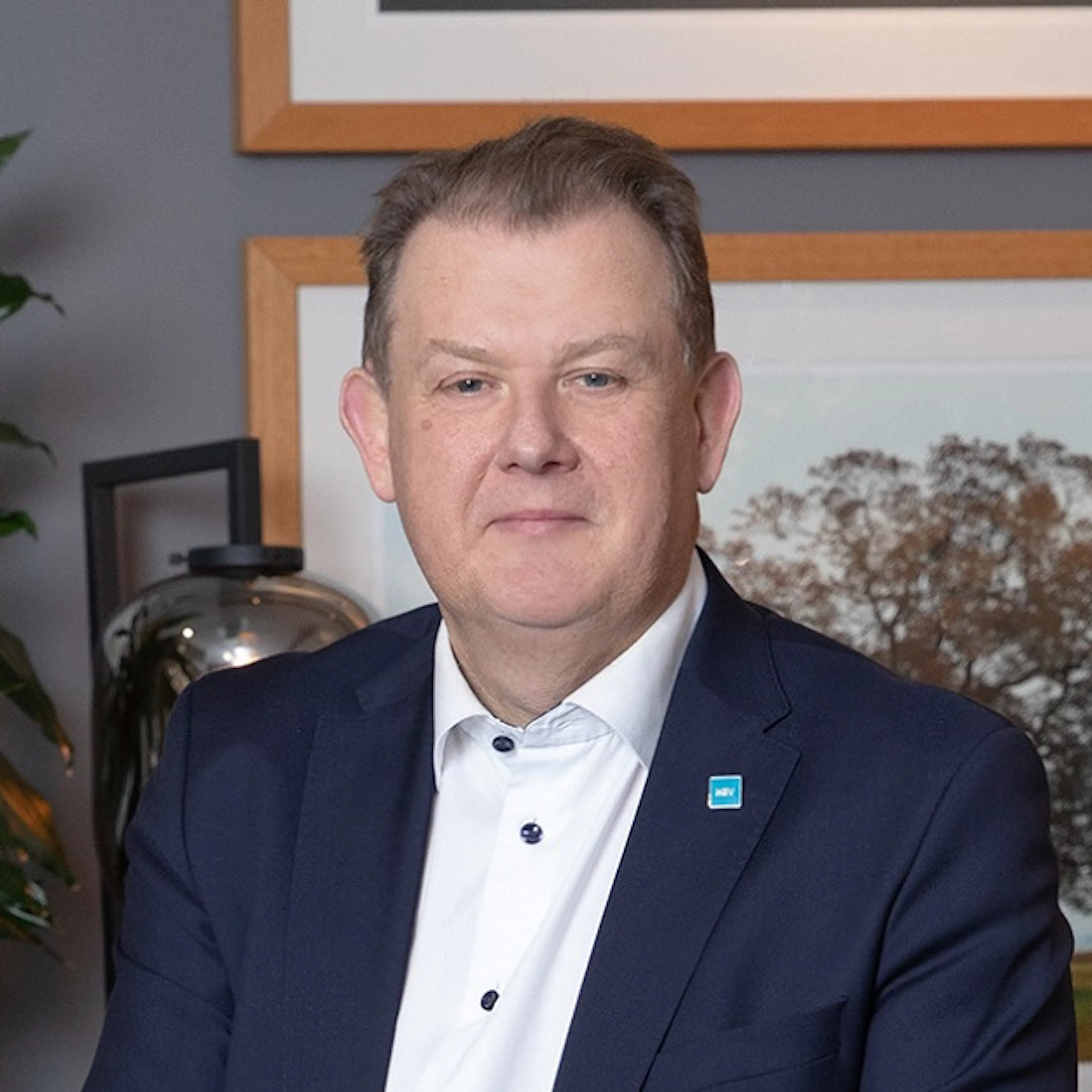 Man i vit skjorta och blå kostym. Sitter framför ett fotografi med ett träd på.