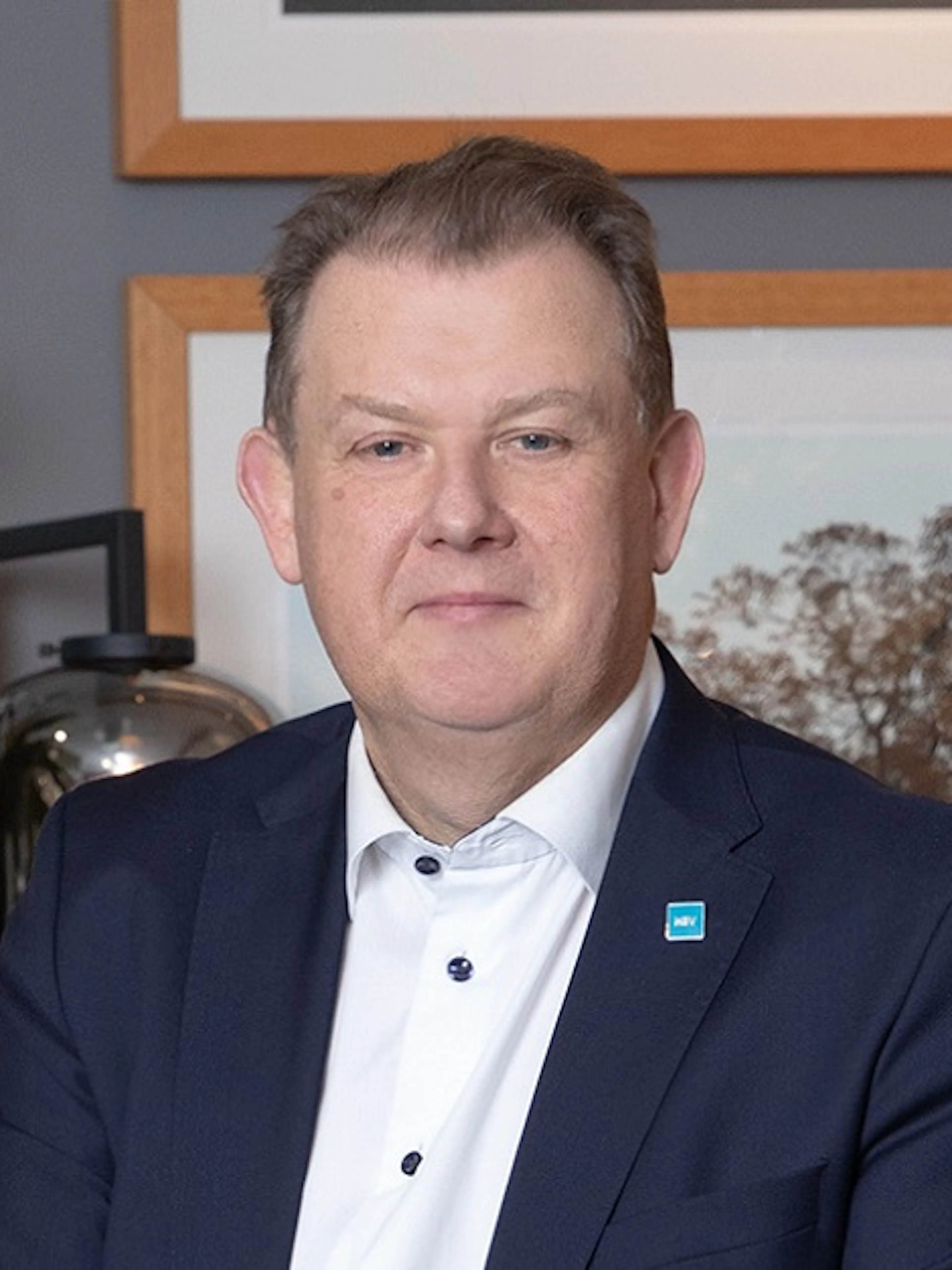 Man i vit skjorta och blå kostym. Sitter framför ett fotografi med ett träd på.