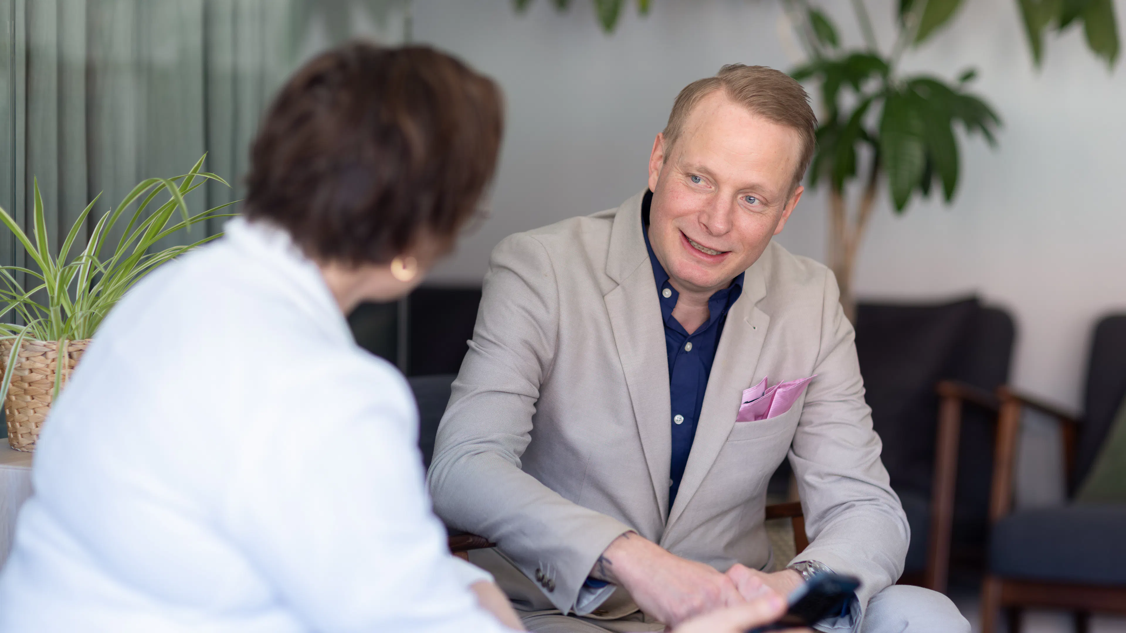 Man i ljus kostym och blå skjorta sitter ner och pratar med kvinna i vit kavaj.