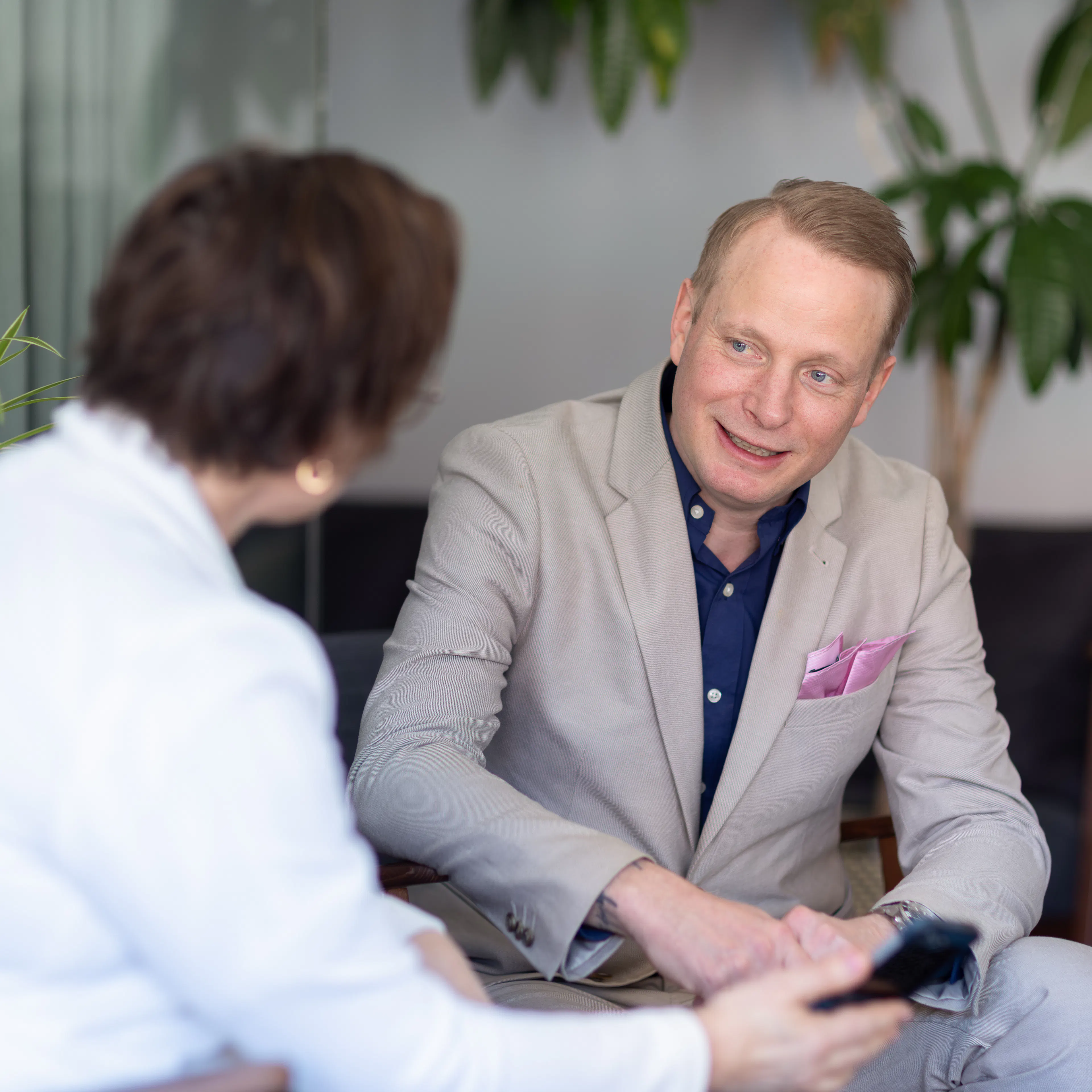 Man i ljus kostym och blå skjorta sitter ner och pratar med kvinna i vit kavaj.