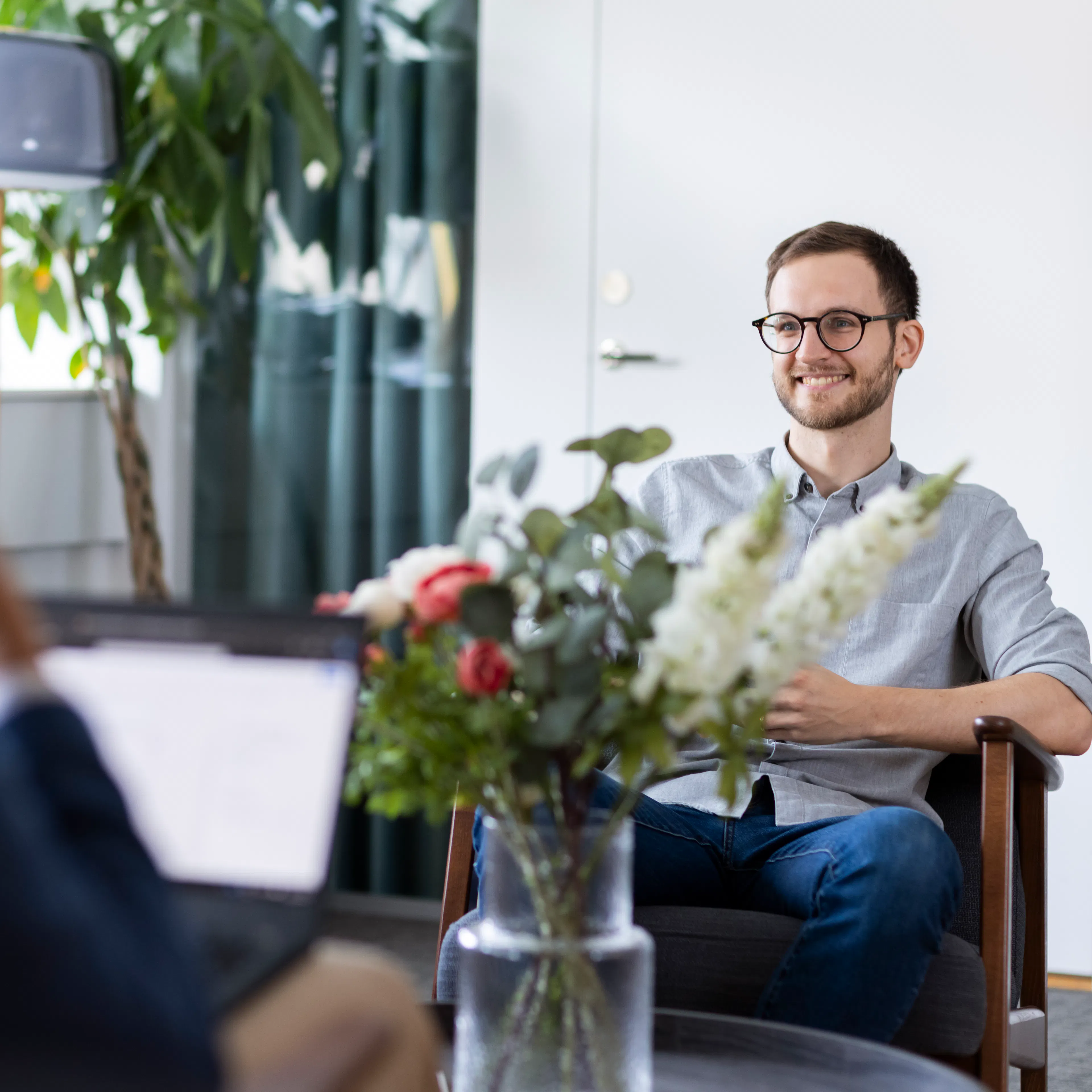 Man i ljus skjorta som sitter i en fotölj bakom en stor blomsterbukett.
