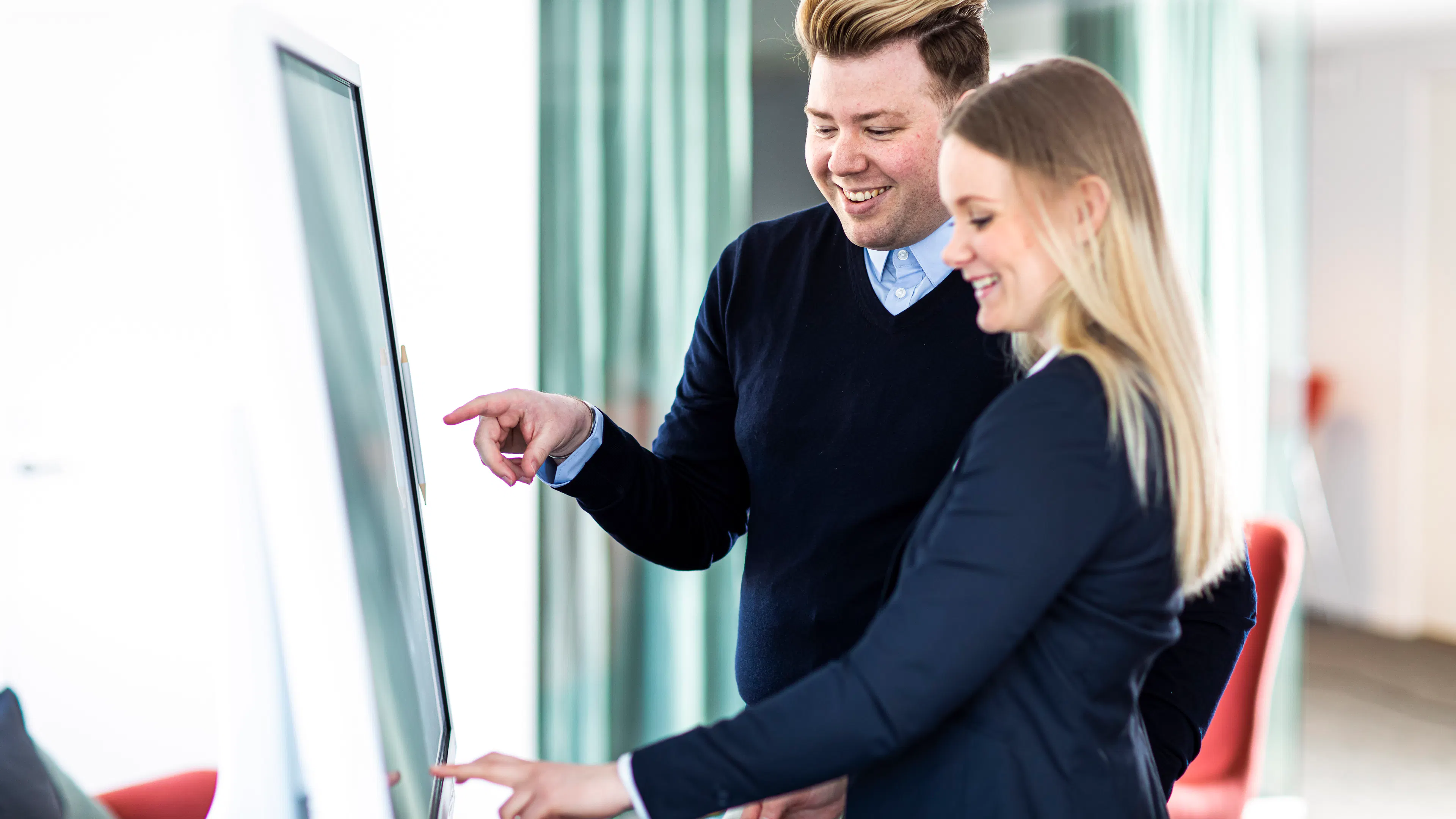 Ein Mann in einem dunklen Pullover und eine Frau in einem schwarzen Kostüm, die vor einem digitalen Bildschirm stehen