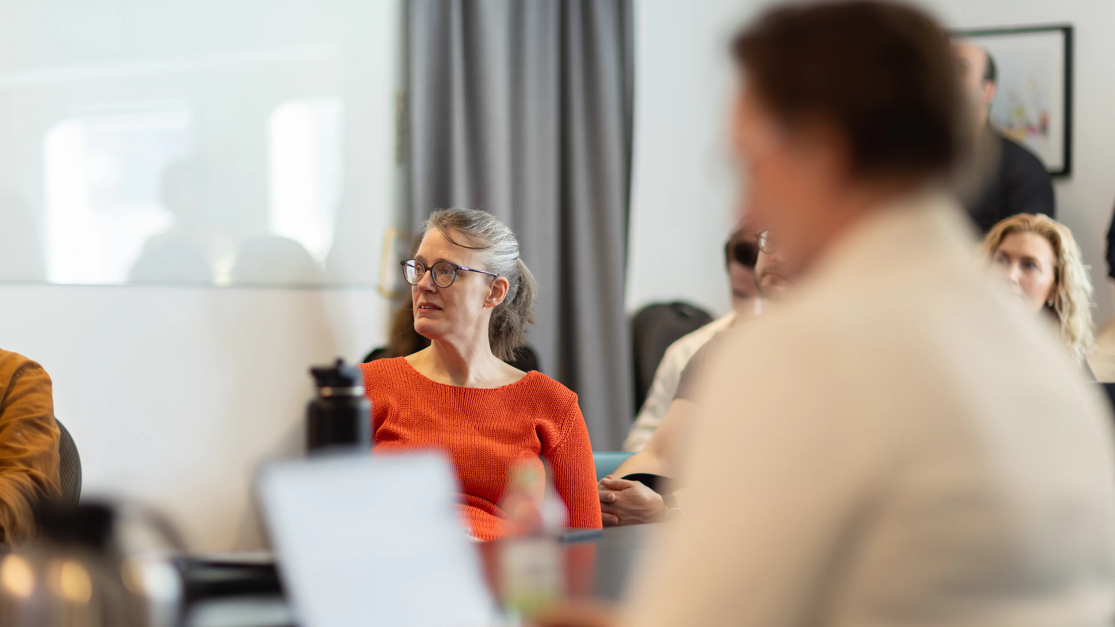 Kvinna i röd tröja och glasögon i fokus. Kvinna i ljus kavaj i förgrund som tittar på en skärm.