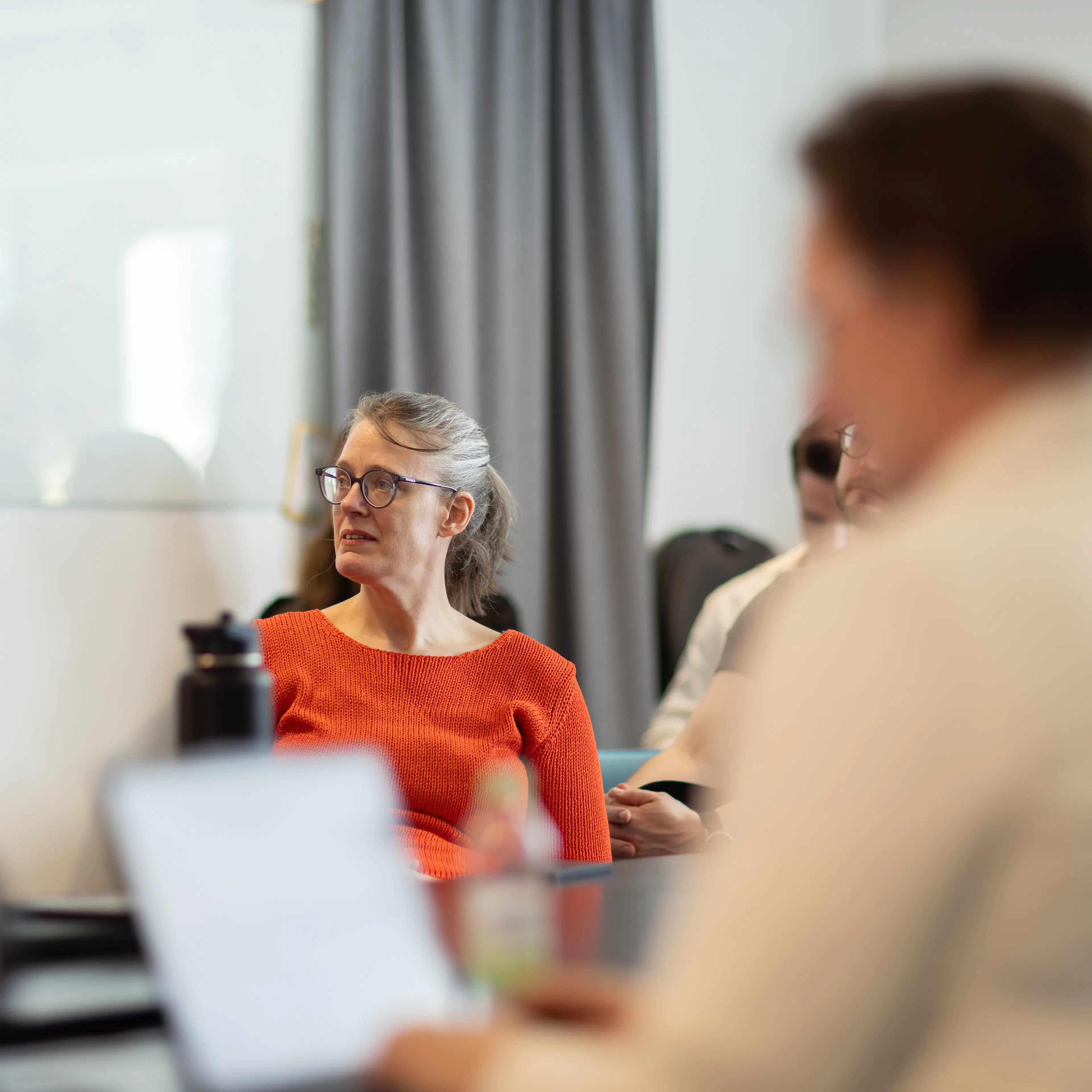 Kvinna i röd tröja och glasögon i fokus. Kvinna i ljus kavaj i förgrund som tittar på en skärm.