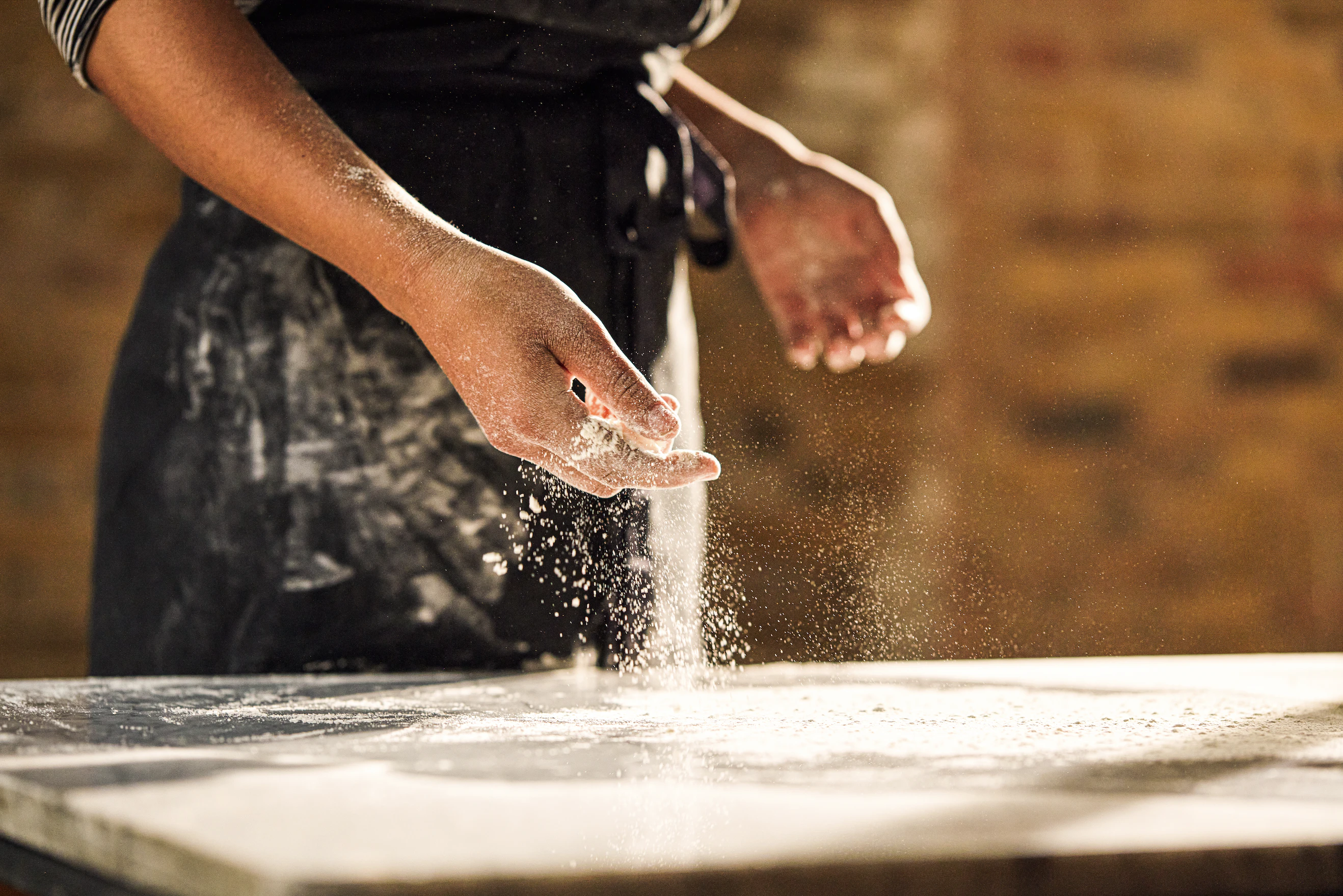 A person in a blue apron scattering flour over a marble surface.