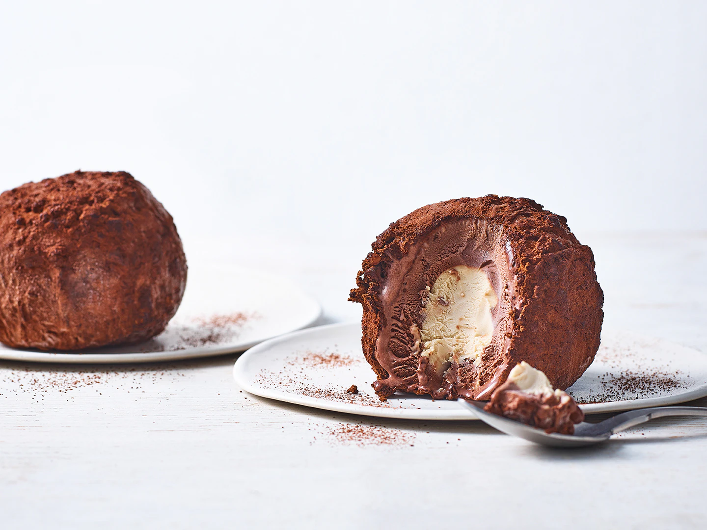 Two chocolate and hazelnut tartufi on white plates, one has a quarter cut out from it. A spoonful of the gelato sits by a plate.