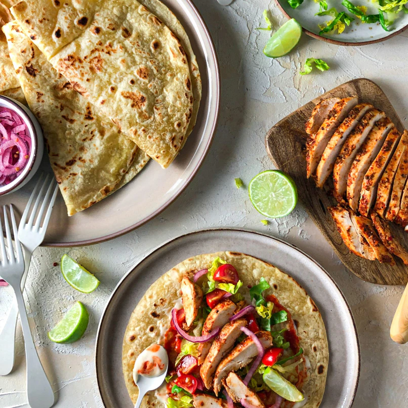 Shot from above, three plates are in view. One has folded Piadina on it, with a small pot of pickled red onions, while another has an assembled lime and chilli chicken piadina. The third plate has sliced chicken breast on it, wedges of lime are scattered between the plates.