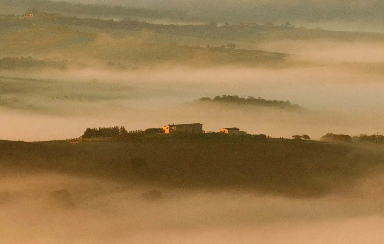 Rolling hills and fog surround a small collection of buildings on top of a hill.