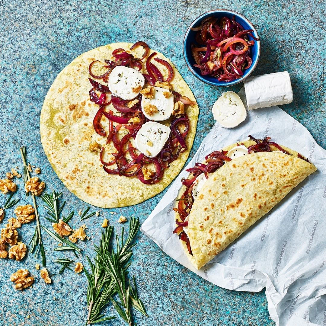 Two Paidian flatbreads are filled with red onions, goats cheese and walnuts, one is open one is folded over. A ramekin of caramelised red onions is next to a log of goats cheese.