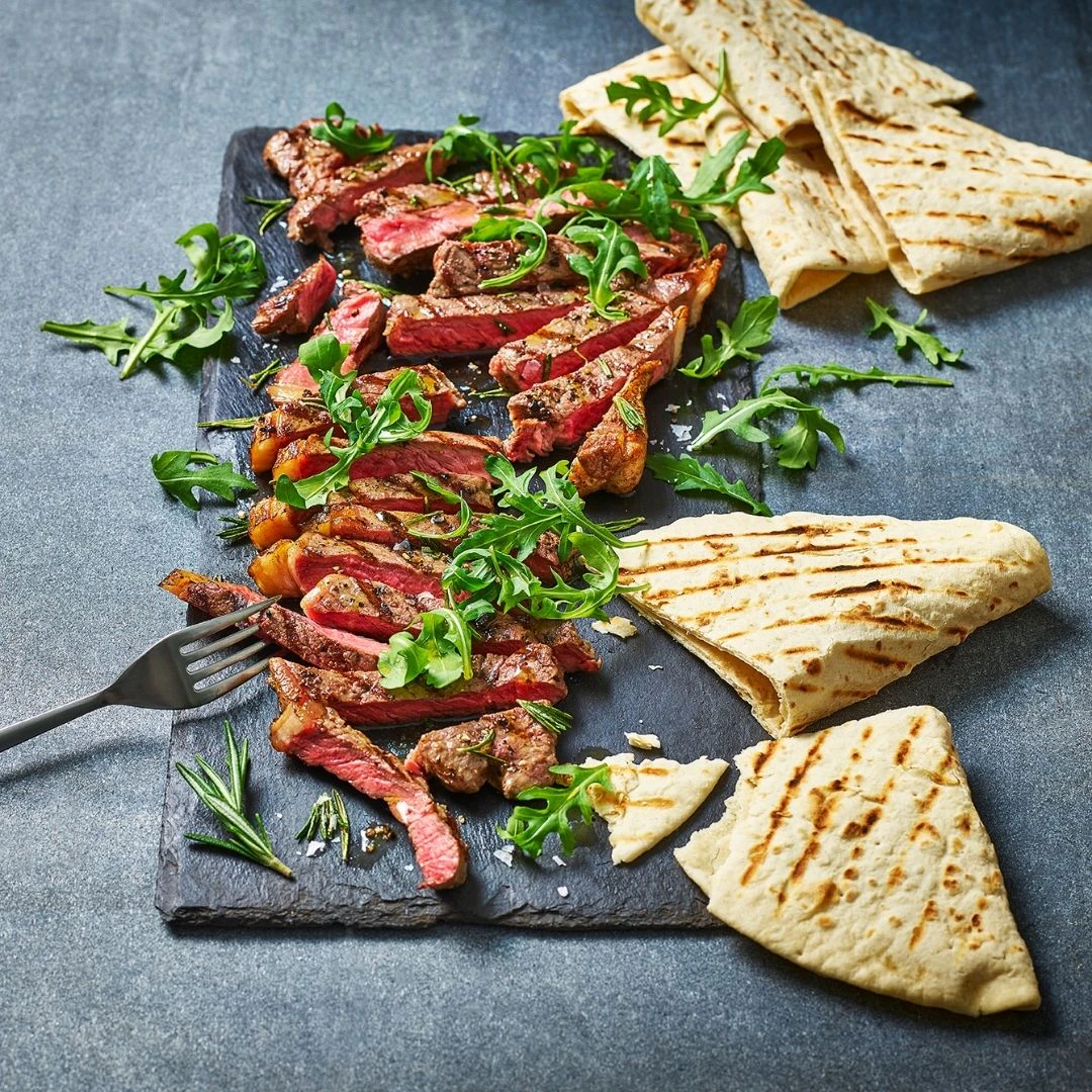 Slices of steak are on a slate board with rocket leaves scattered over them. To the side are charred slices of Piadina Emiliana.