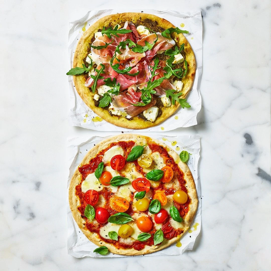 Two pizzas shot overhead, one is topped with Pesto, Prosciutto & Goats Cheese, the other with tomatoes, mozzarella and fresh basil.