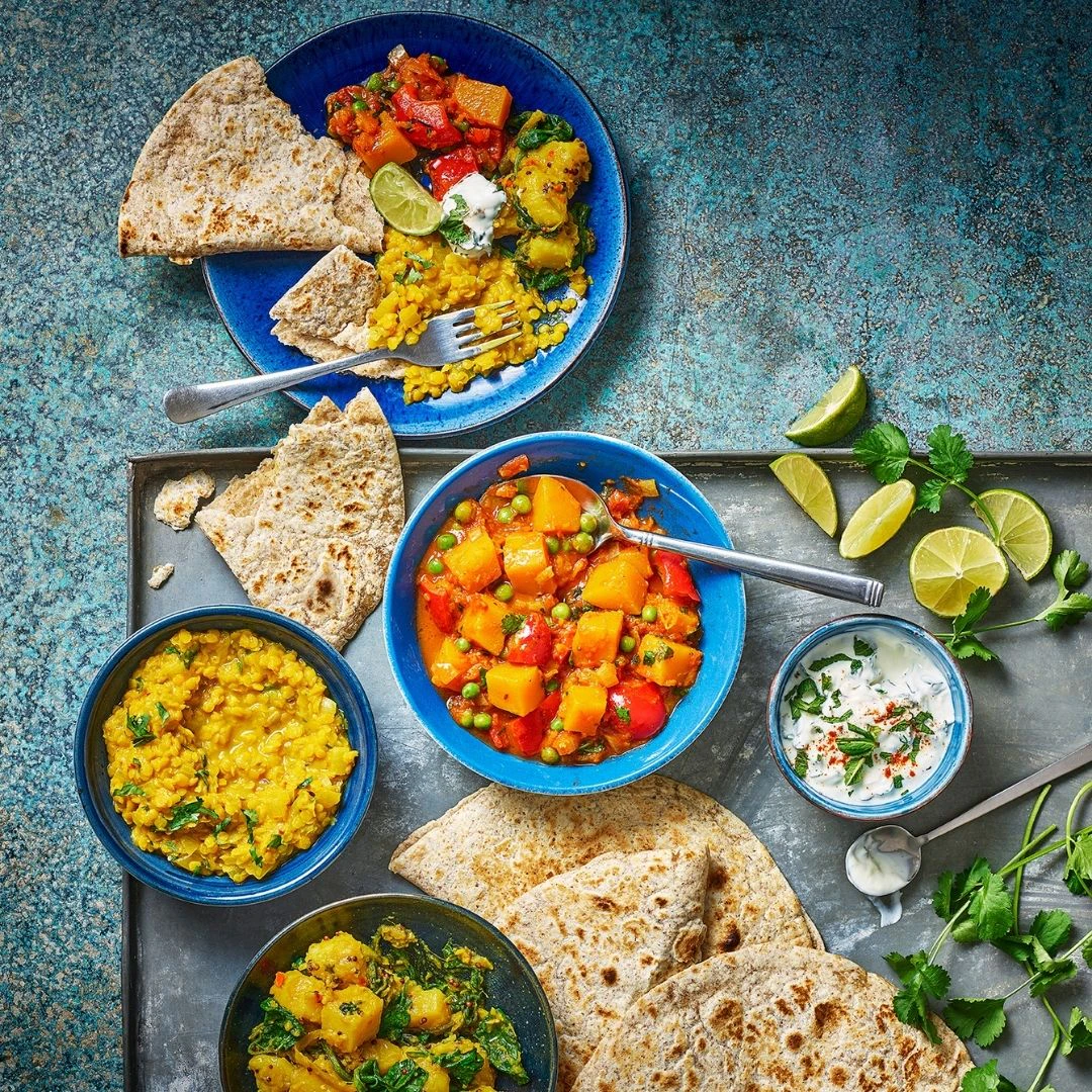 An overhead shot of four bowls of curry, with Wholeblend Piadina, wedges of lime, and fresh coriander scattered between the bowls.