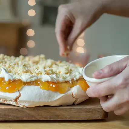 A hand is sprinkling crushed amaretti biscuits over a pavlova.