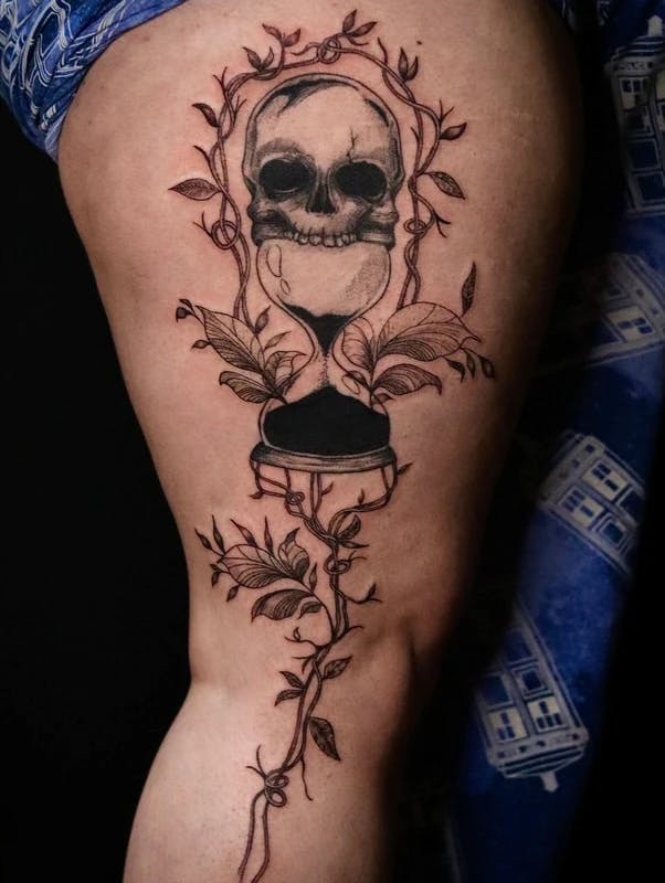 Tattoo of a skull with an hourglass coming out of its mouth, surrounded by vines.