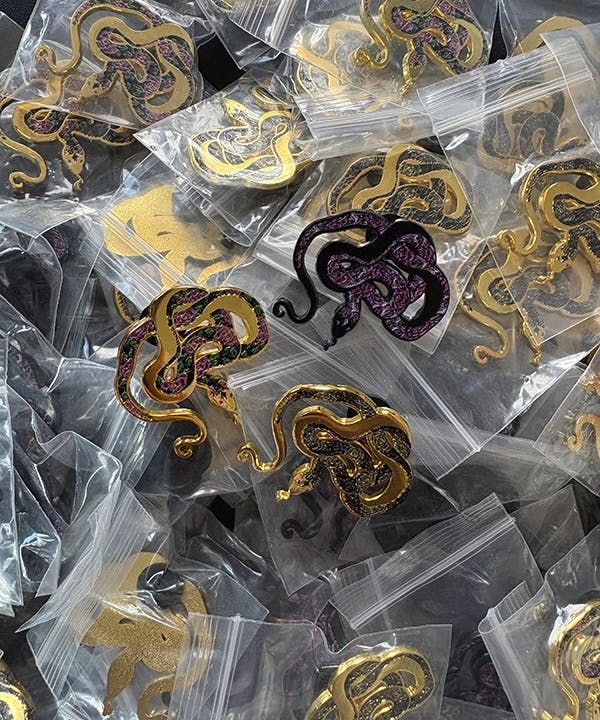 Three versions of Crown & Serpent's logo pins on top of a pile of packaged pins.