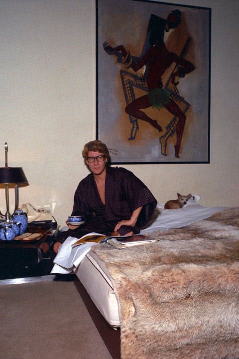 Saint Laurent in his rue de Babylone bedroom in 1974, with Hazel, his Chihuahua. Behind the bed hangs a Pierre Colin study of Josephine Baker; a lynx spread covers the bed.