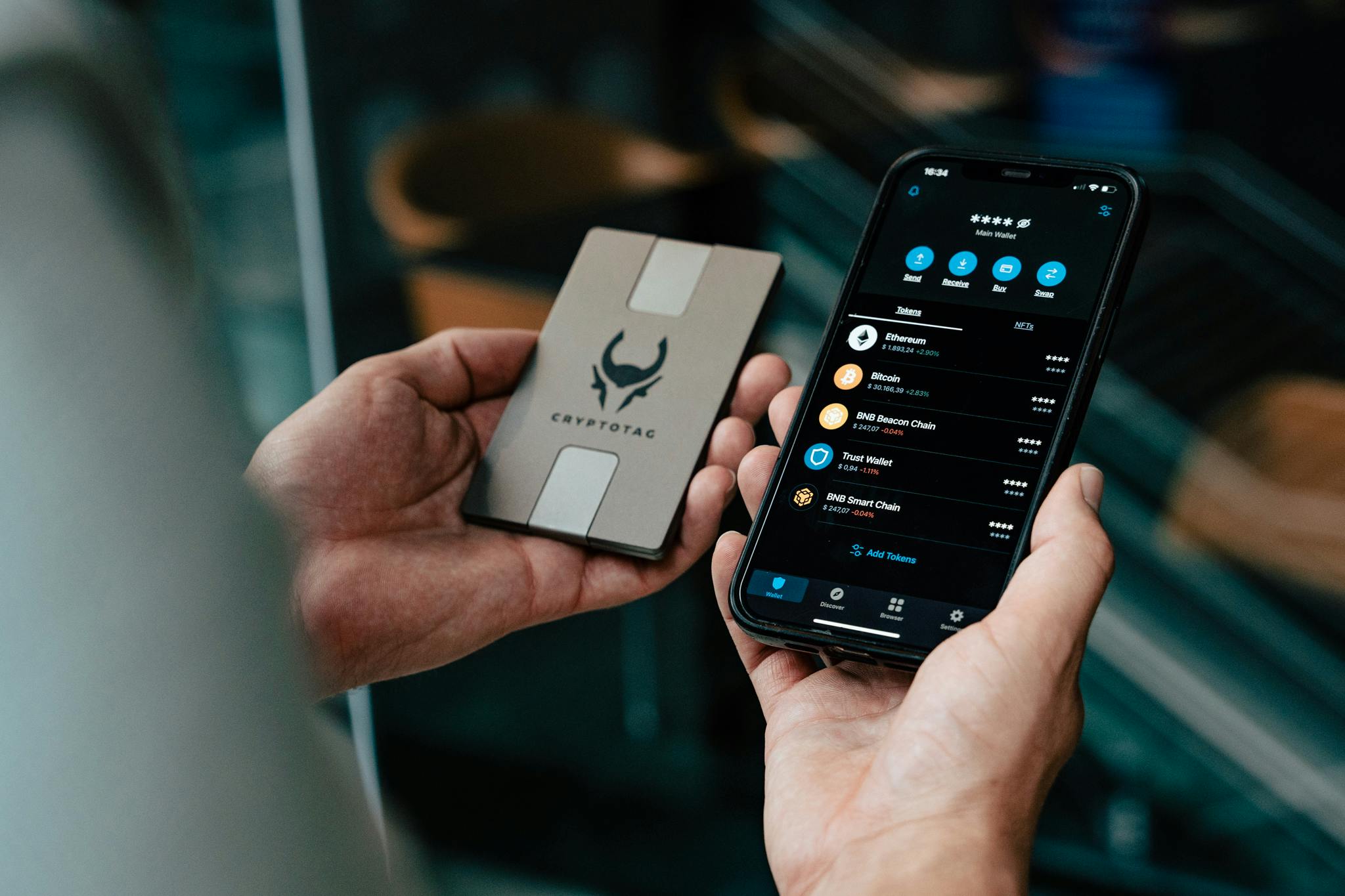 Two hands holding an iPhone with the Trust Wallet app opened, in the other hand a titanium Cryptotag