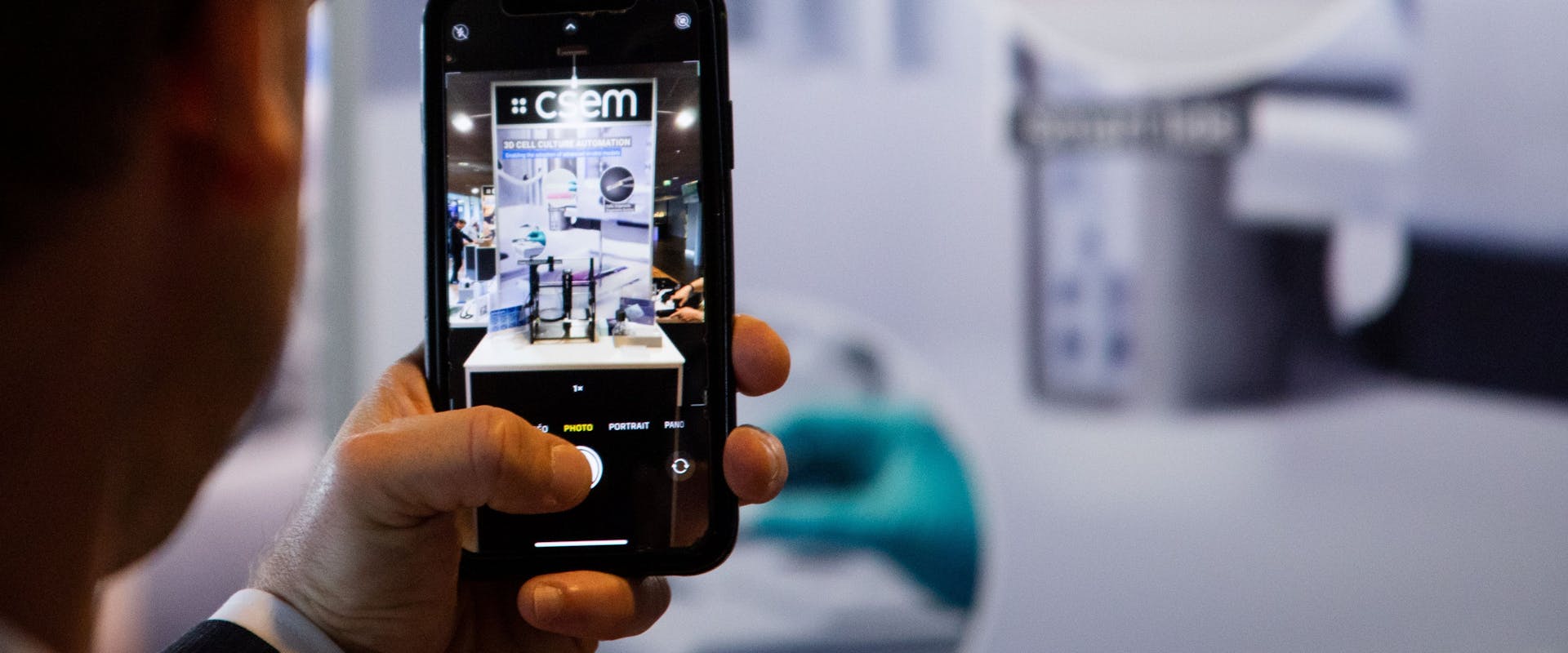 A man seen from the back, holding a smartphone with a picture of a CSEM booth