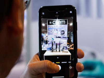 A man seen from the back, holding a smartphone with a picture of a CSEM booth