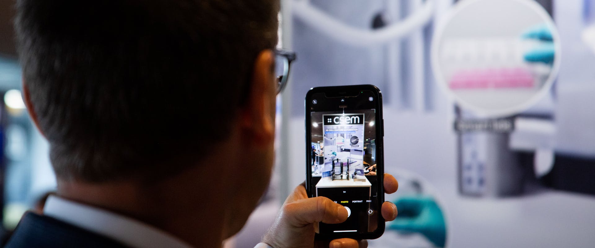 A man seen from the back, holding a smartphone with a picture of a CSEM booth
