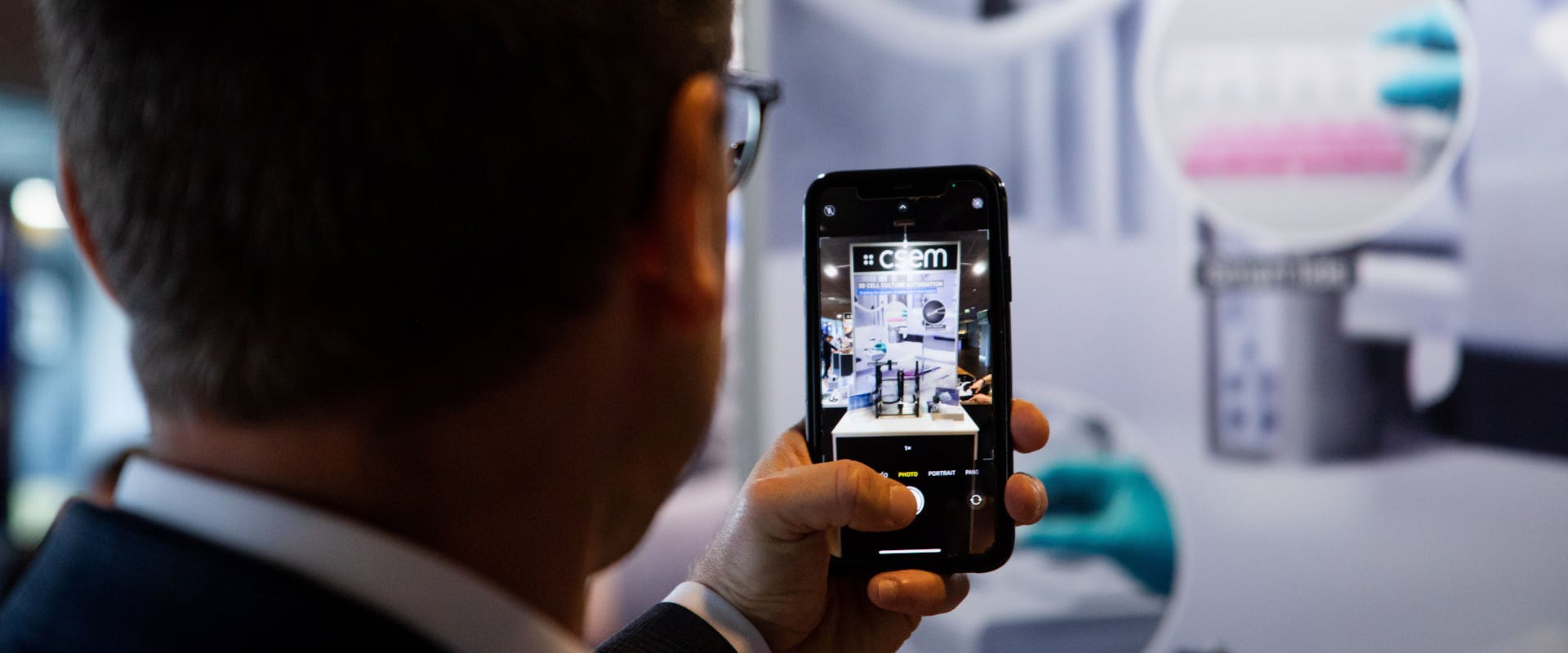 A man seen from the back, holding a smartphone with a picture of a CSEM booth
