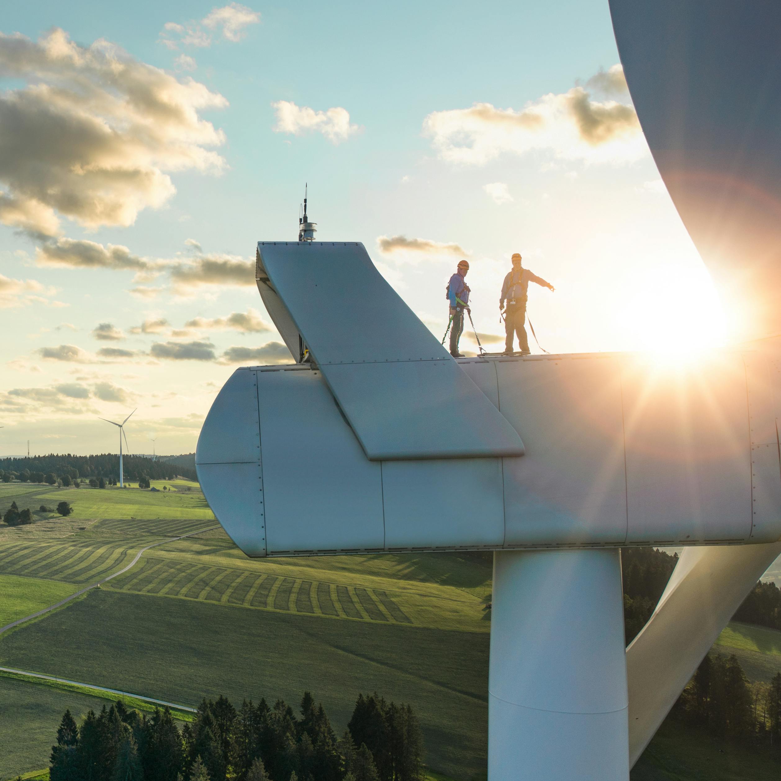 Two people standing on top of a wind turbine, green landscape.
