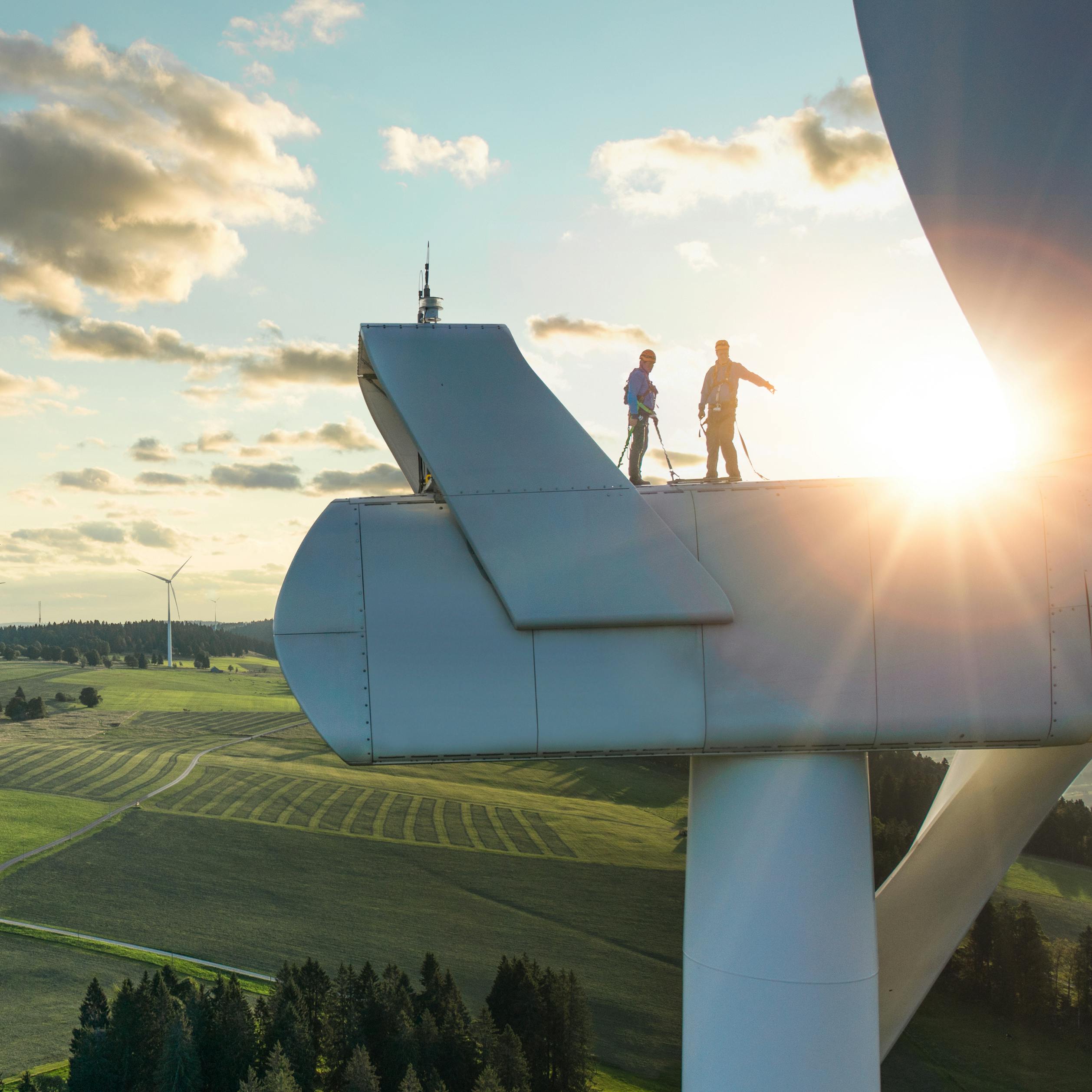 Zwei Menschen stehen auf einer Windkraftanlage, grüne Landschaft.
