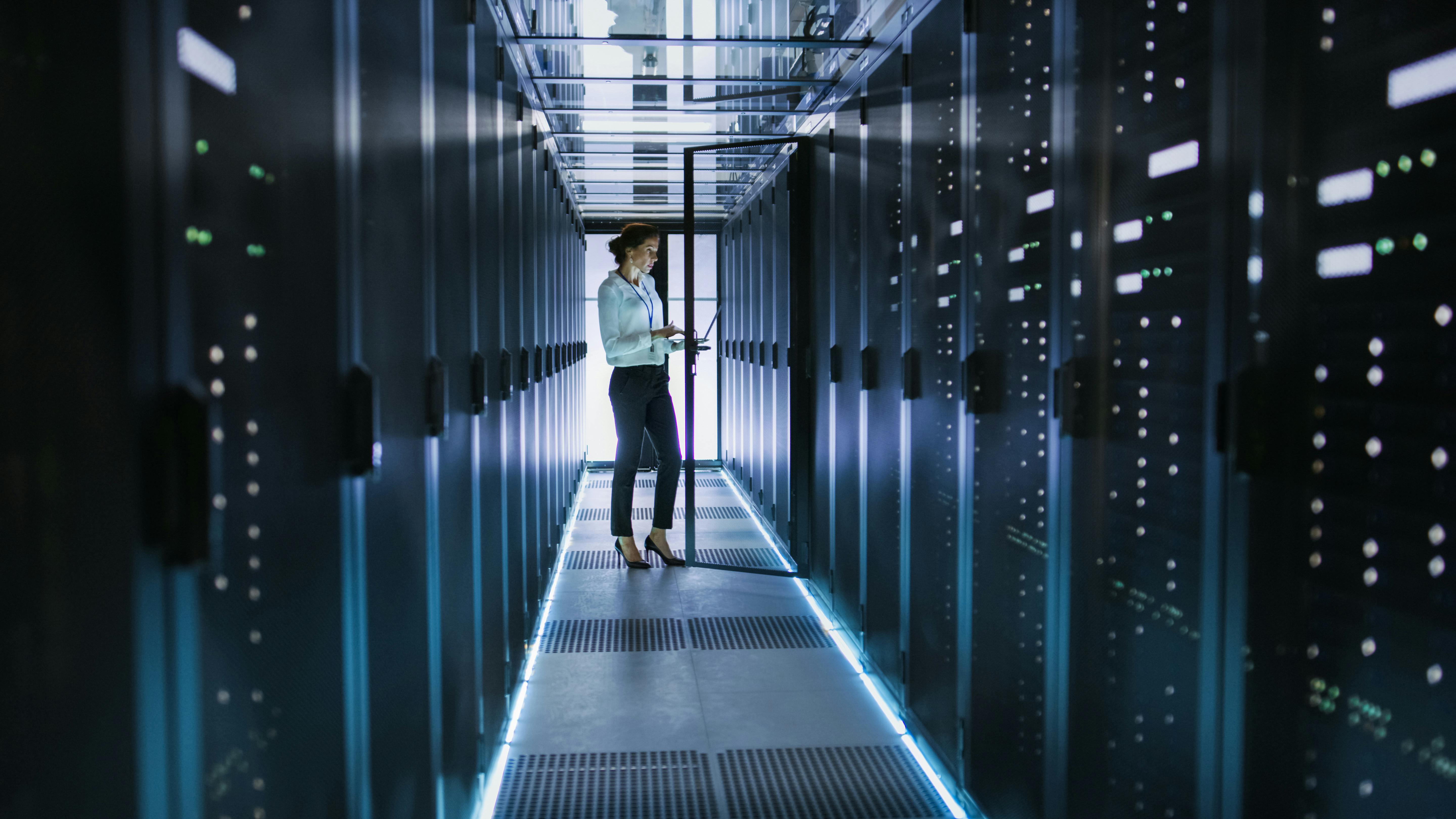 A female office worker standing in a data center server room while holding her computer. 
