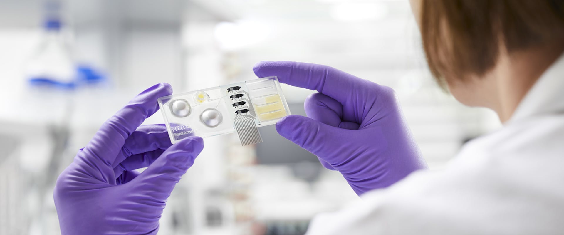 Picture of a woman in lab coat and gloves holding a device