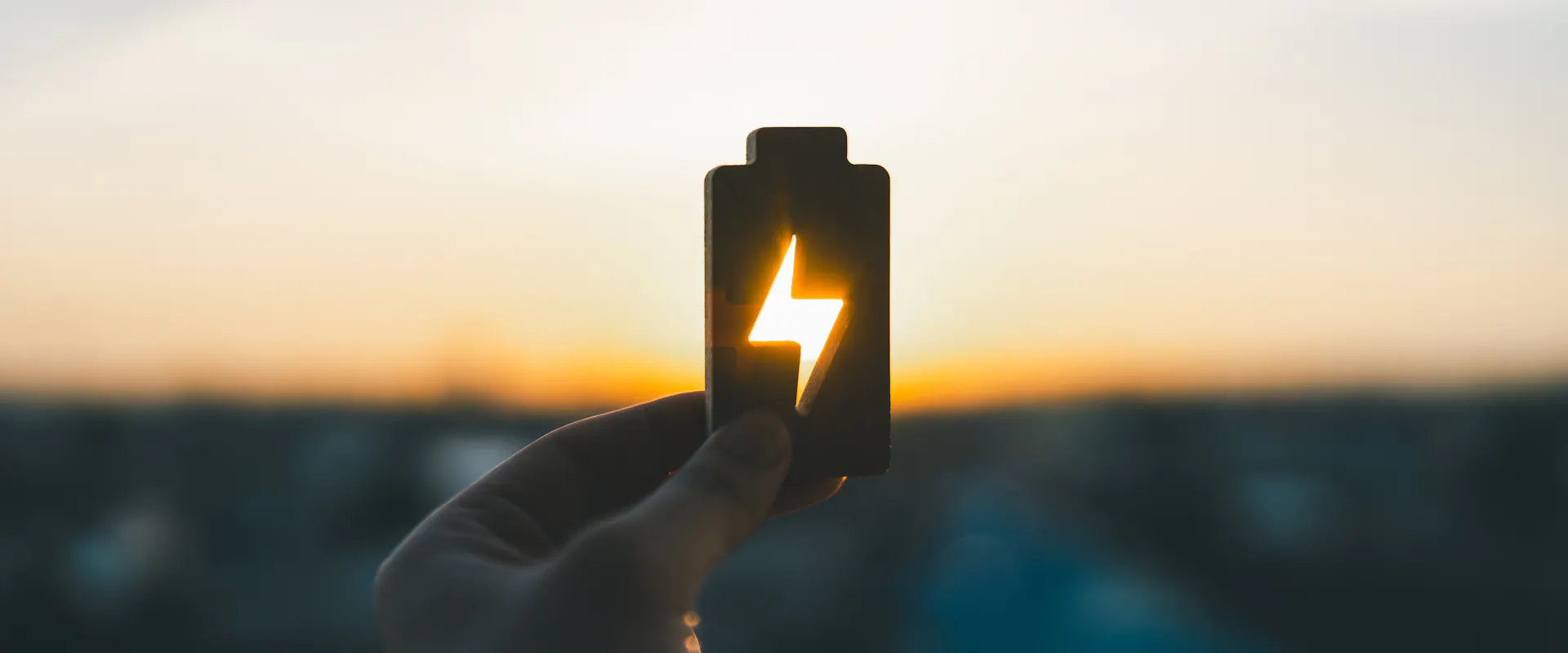 Hand holding battery icon with lightning symbol representing renewable energy and power storage at sunset