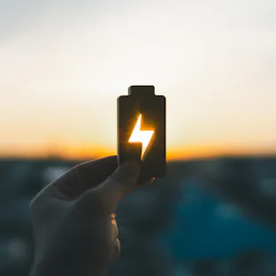 Hand holding battery icon with lightning symbol representing renewable energy and power storage at sunset