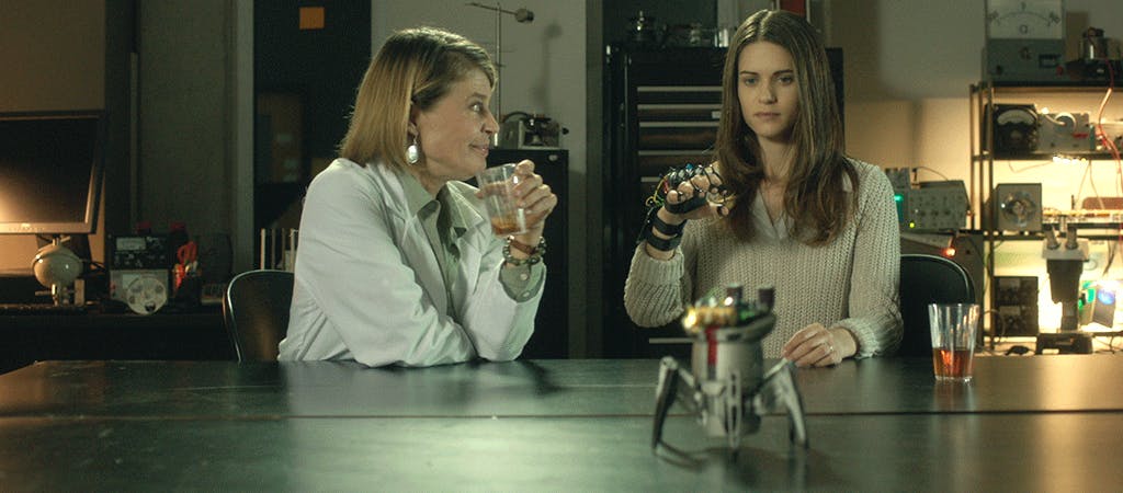two women at table: one holding beverage and one demonstrating a robot