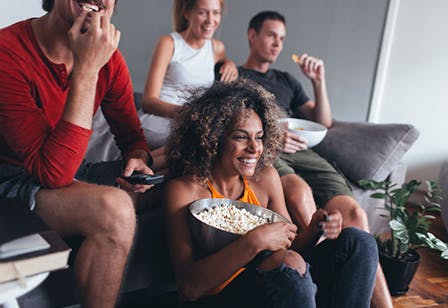 smiling group of people sitting, snacking on chips and popcorn and watching a tv screen