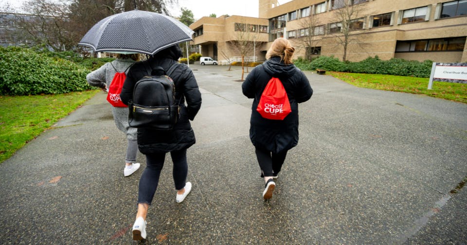 Three people walking across campus, two wearing choose CUPE backpacks, the third carrying an umbrella.