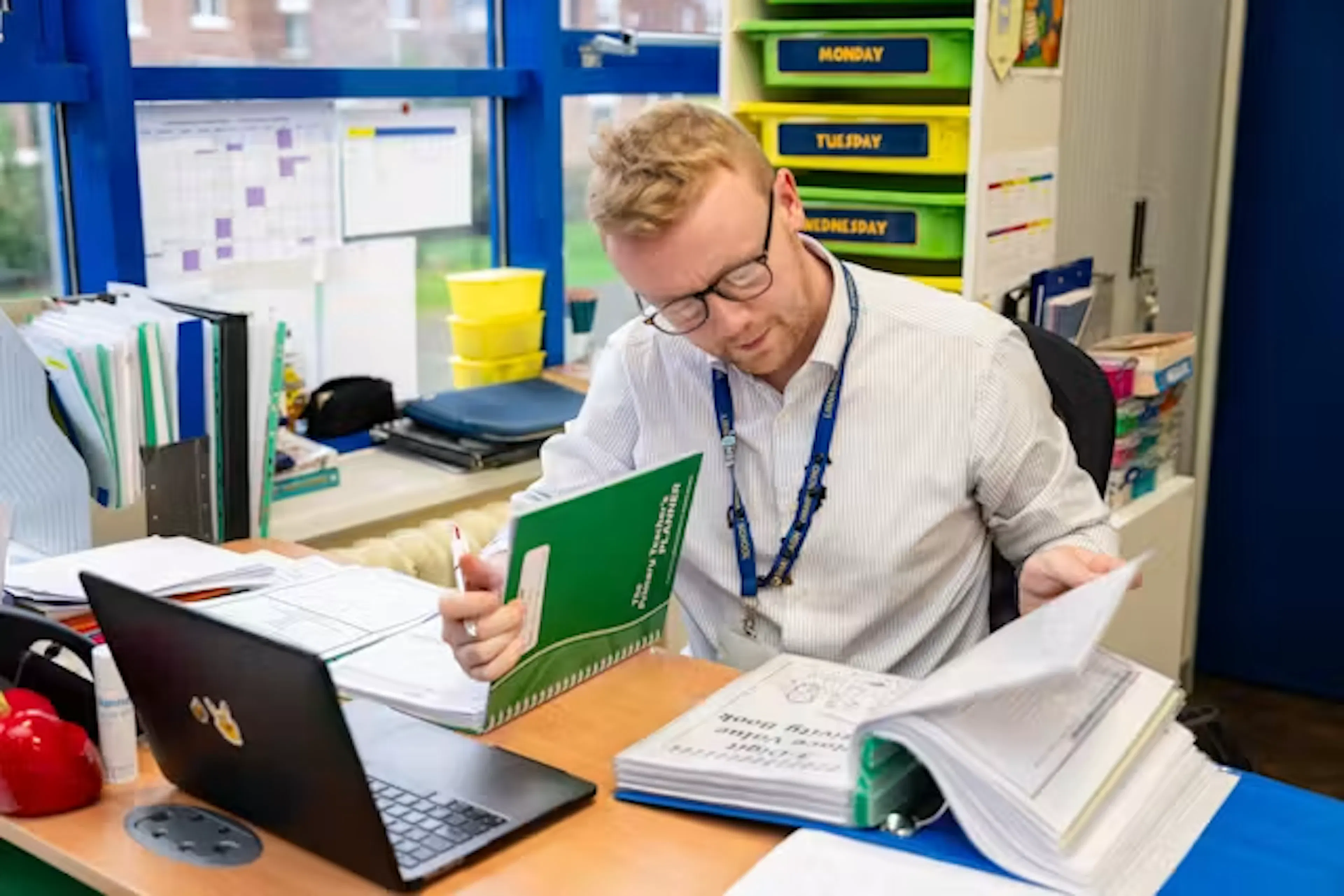A teacher leafing through a file while sitting at a desk with a laptop