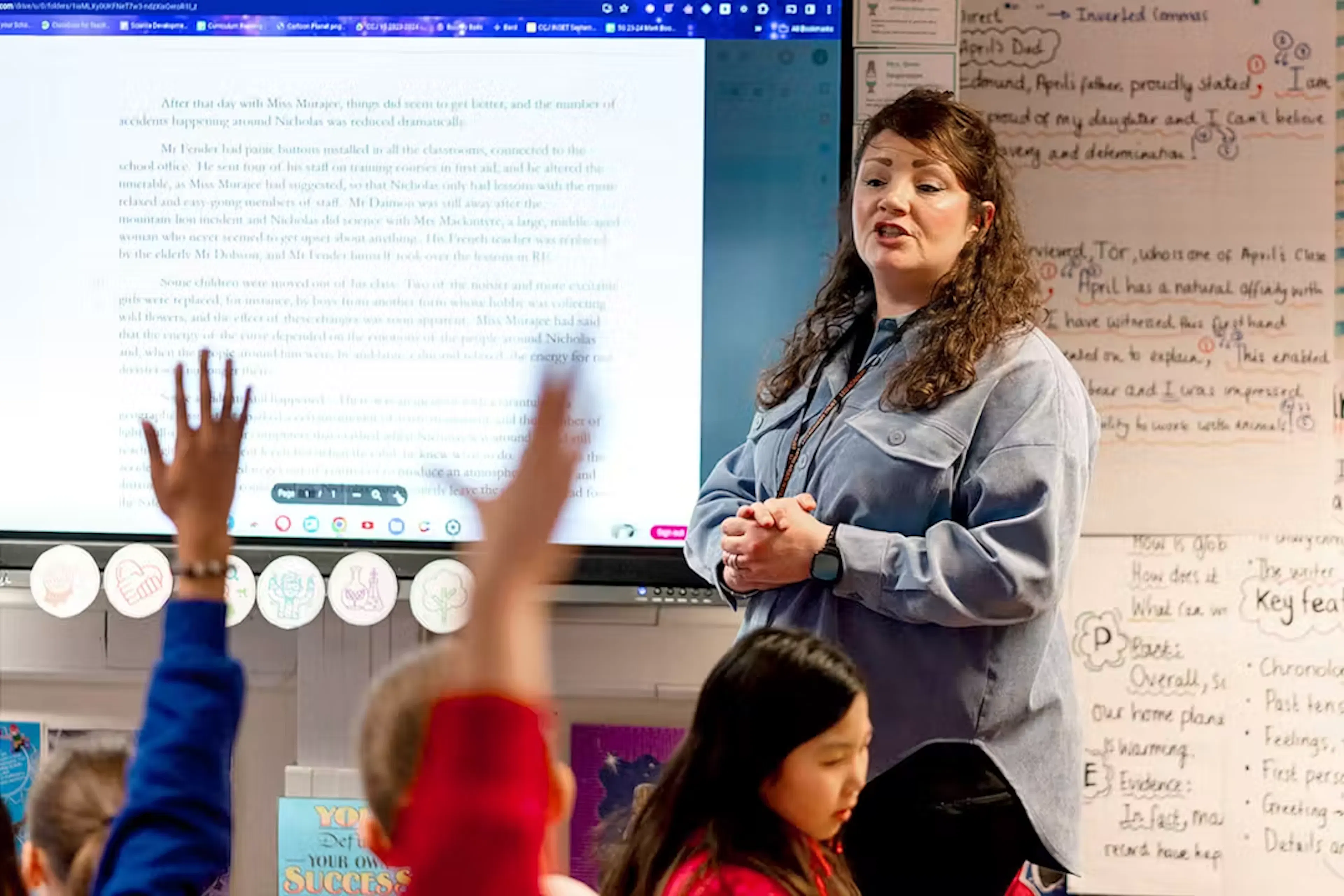 A teacher standing in front of an interactive whiteboard with students raising their hands