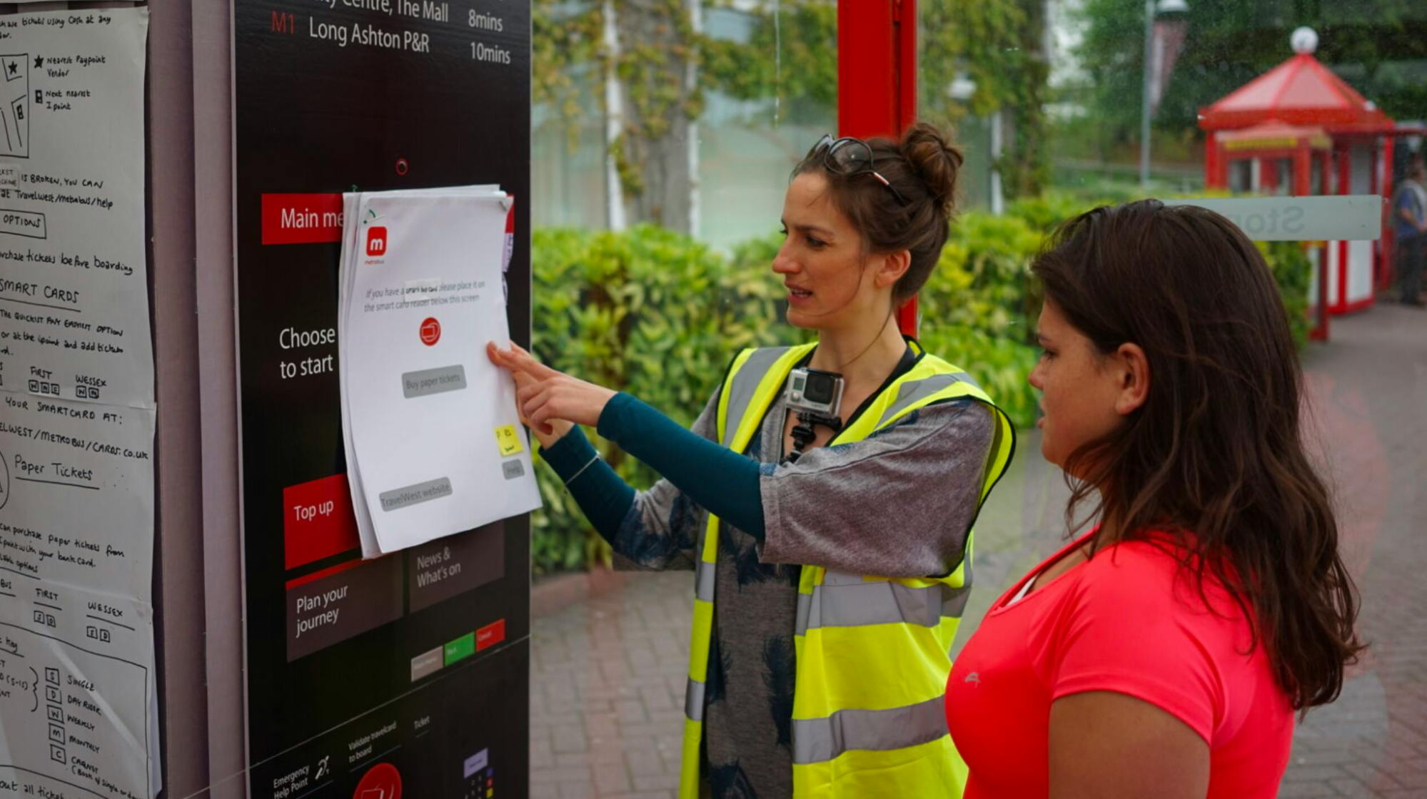 A photo of Anna at a bus stop holding up an A3 piece of paper with ticket purchasing options on, getting feedback from people who use the bus.