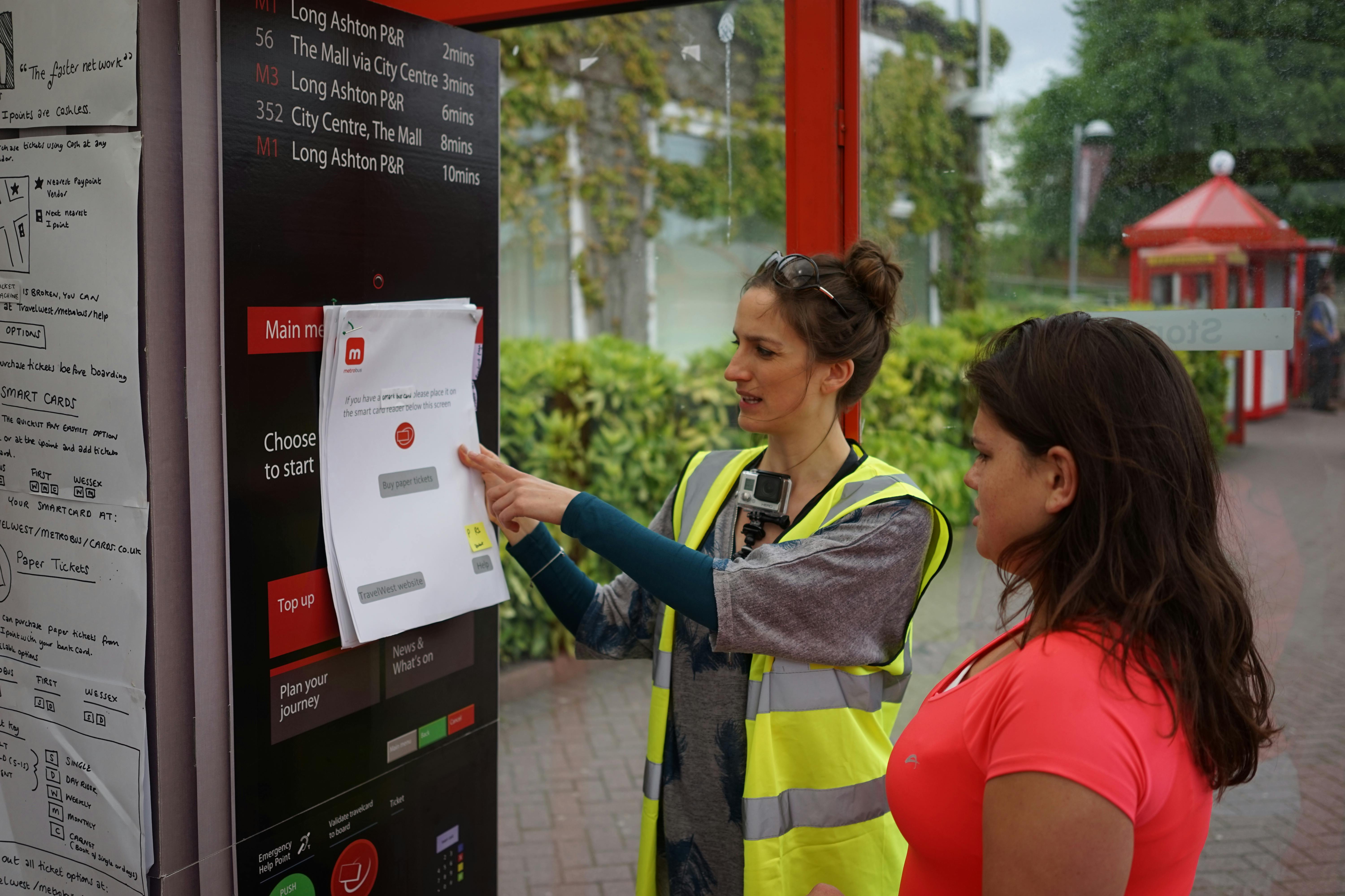 A photo of Anna at a bus stop holding up an A3 piece of paper with ticket purchasing options on, getting feedback from people who use the bus