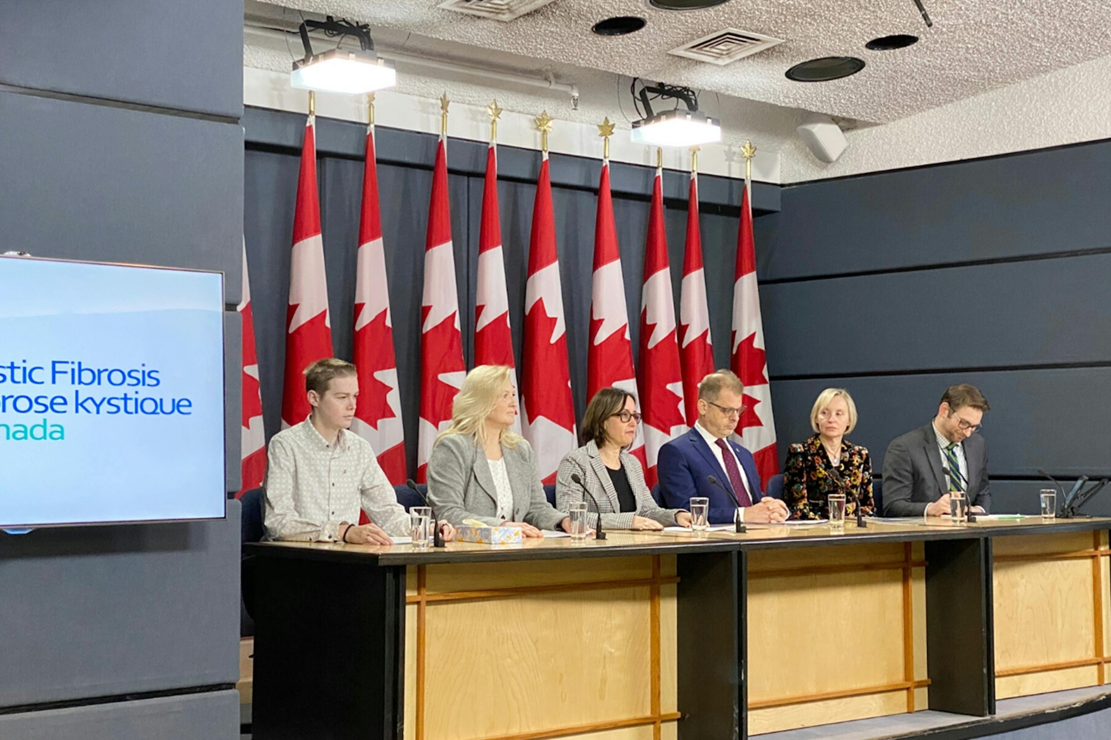 Group of people sitting at desks in front of a line of Canadian flags