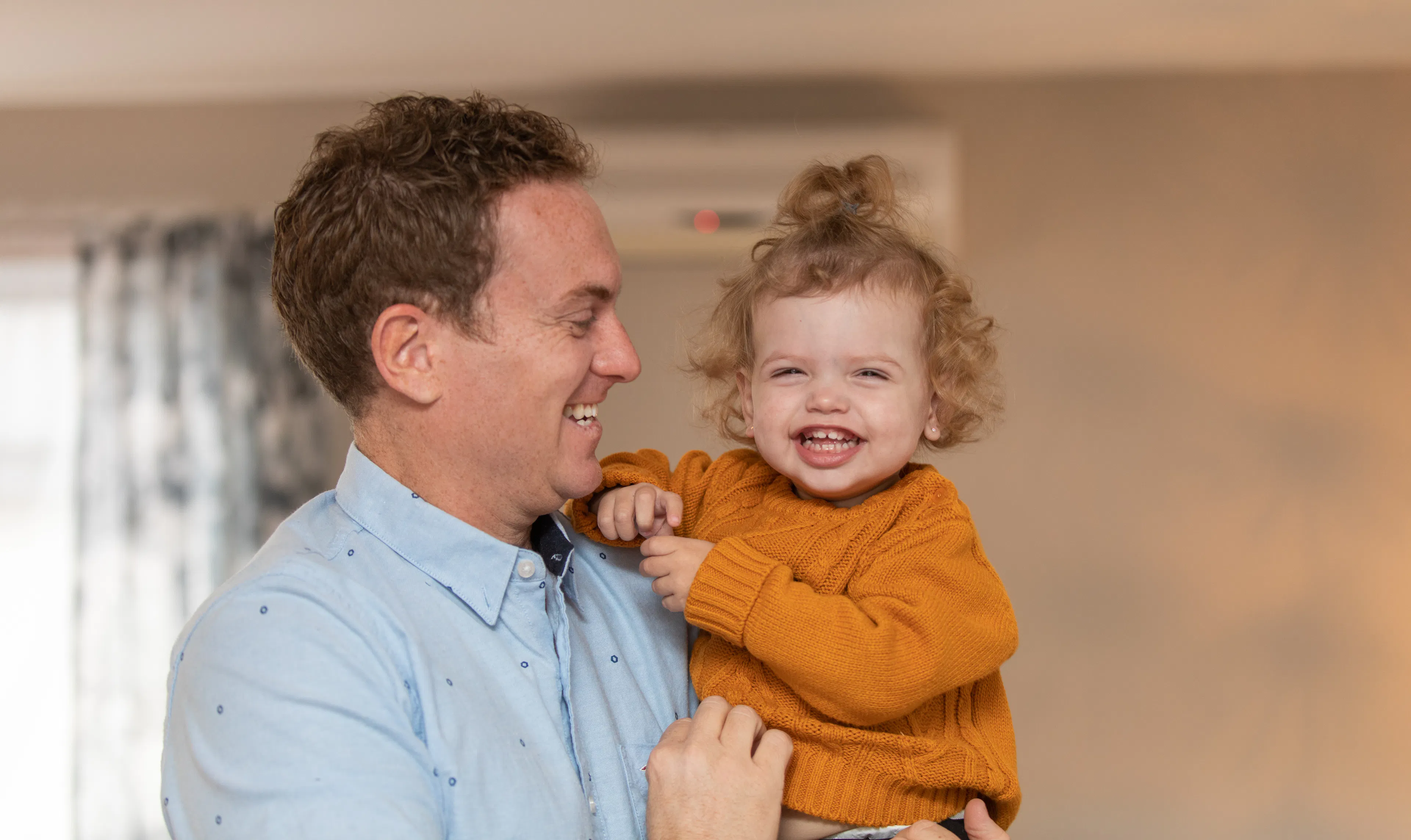 A father holds his young toddler daughter in his arms as she smiles 