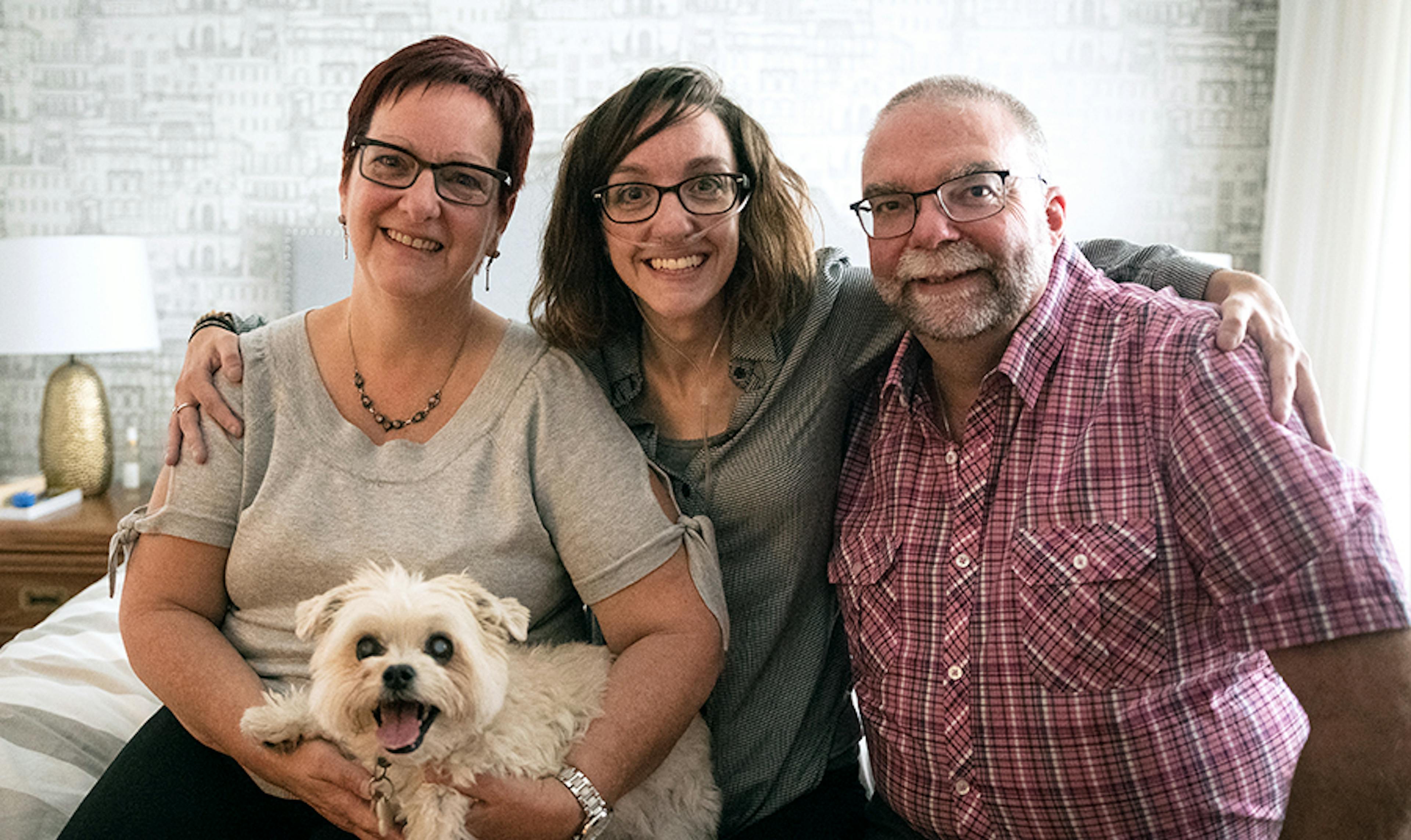 A woman with breathing tube is embraced by her parents