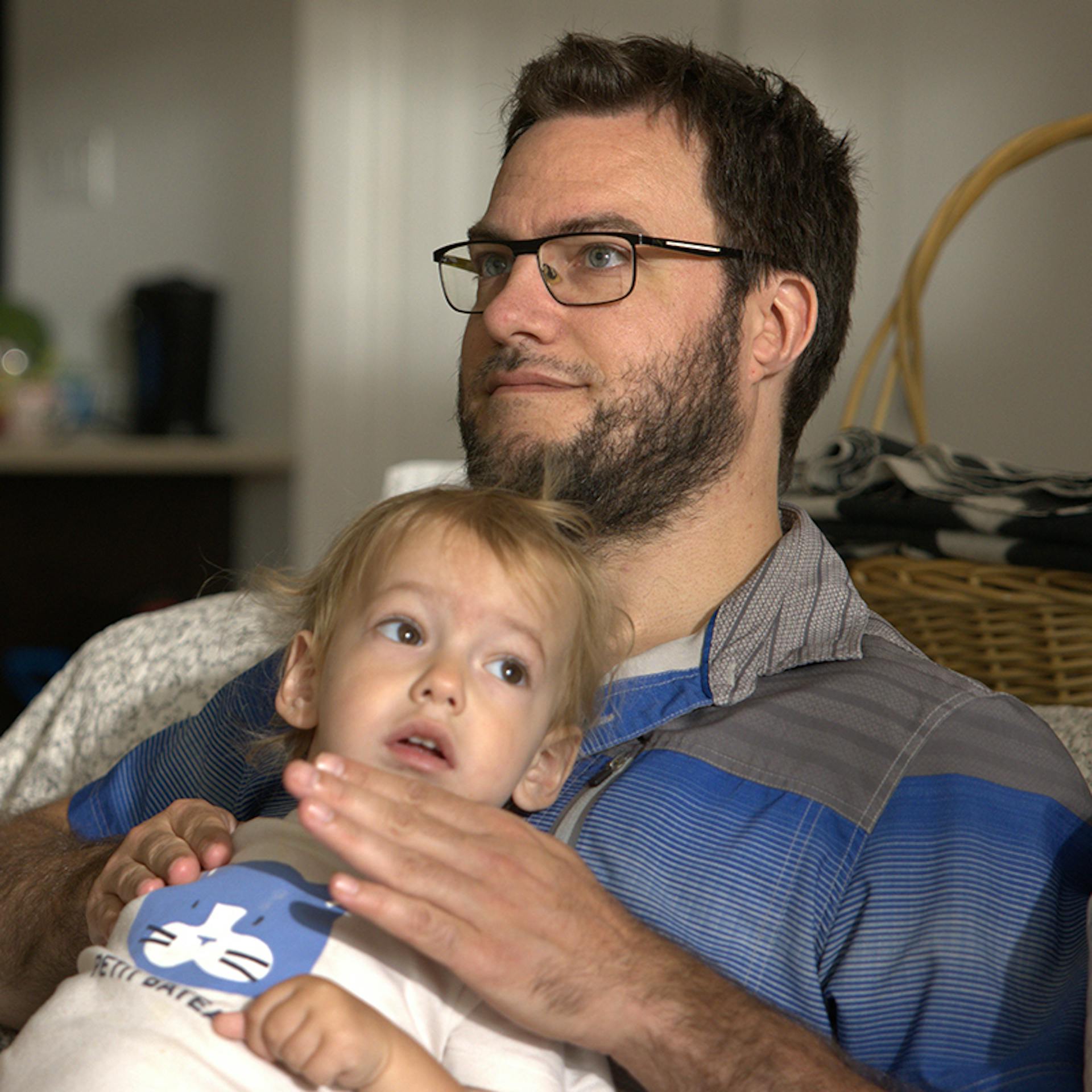 A father sitting on the sofa with his toddler, patting his chest. 