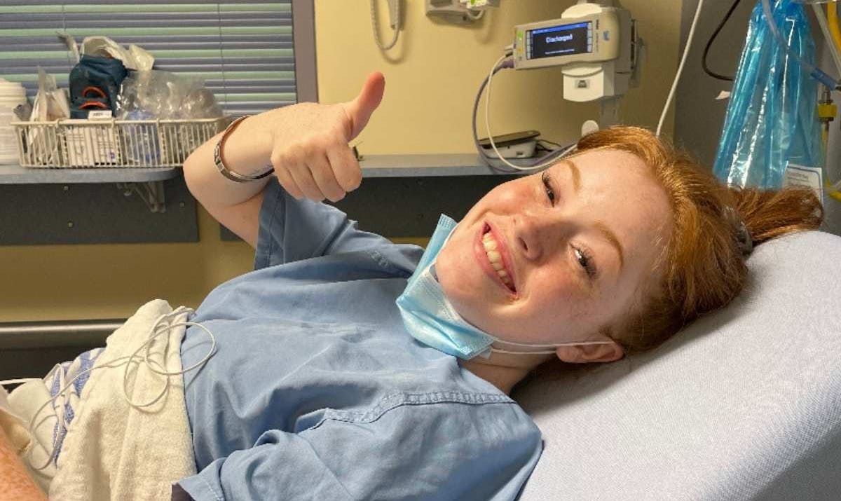 A smiling woman laying on a hospital bed holding a thumbs up.