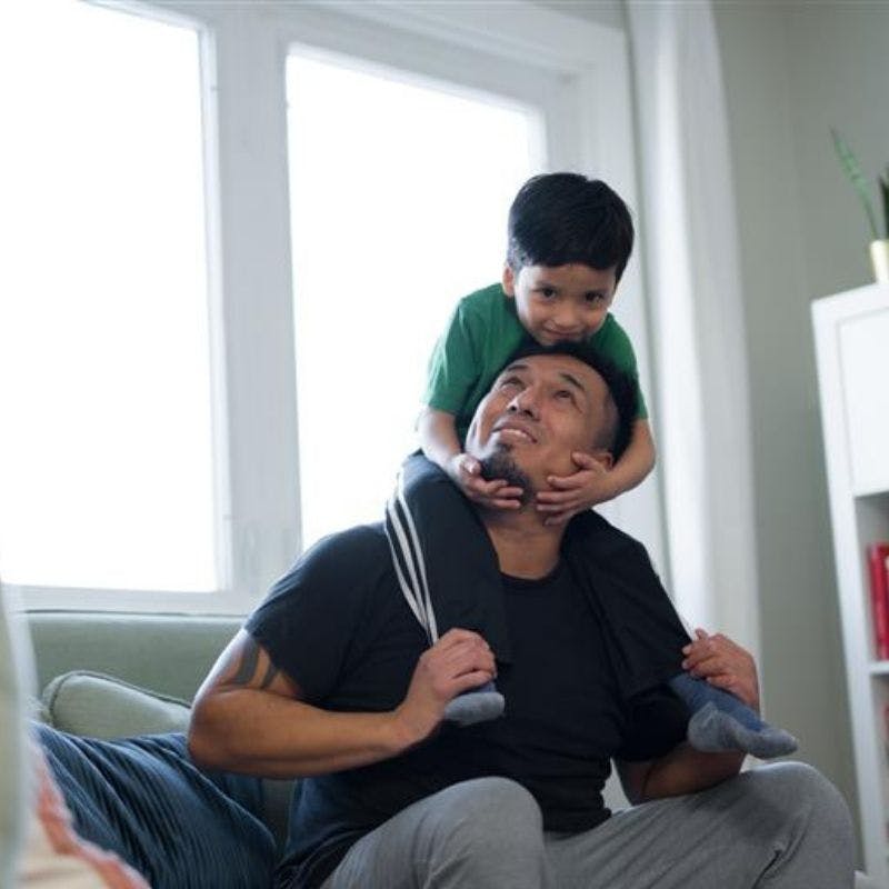 A father and son in a playful post, the son who lives with cystic fibrosis sits on top of the fathers shoulders, the dad looks up at him