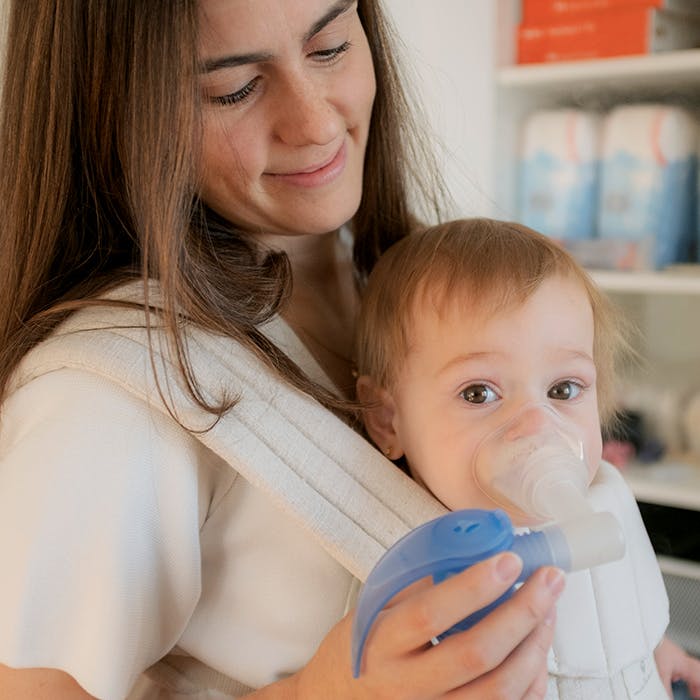 Une jeune maman tient son bébé dans un porte-bébé tout en l'aidant à utiliser son nébuliseur.