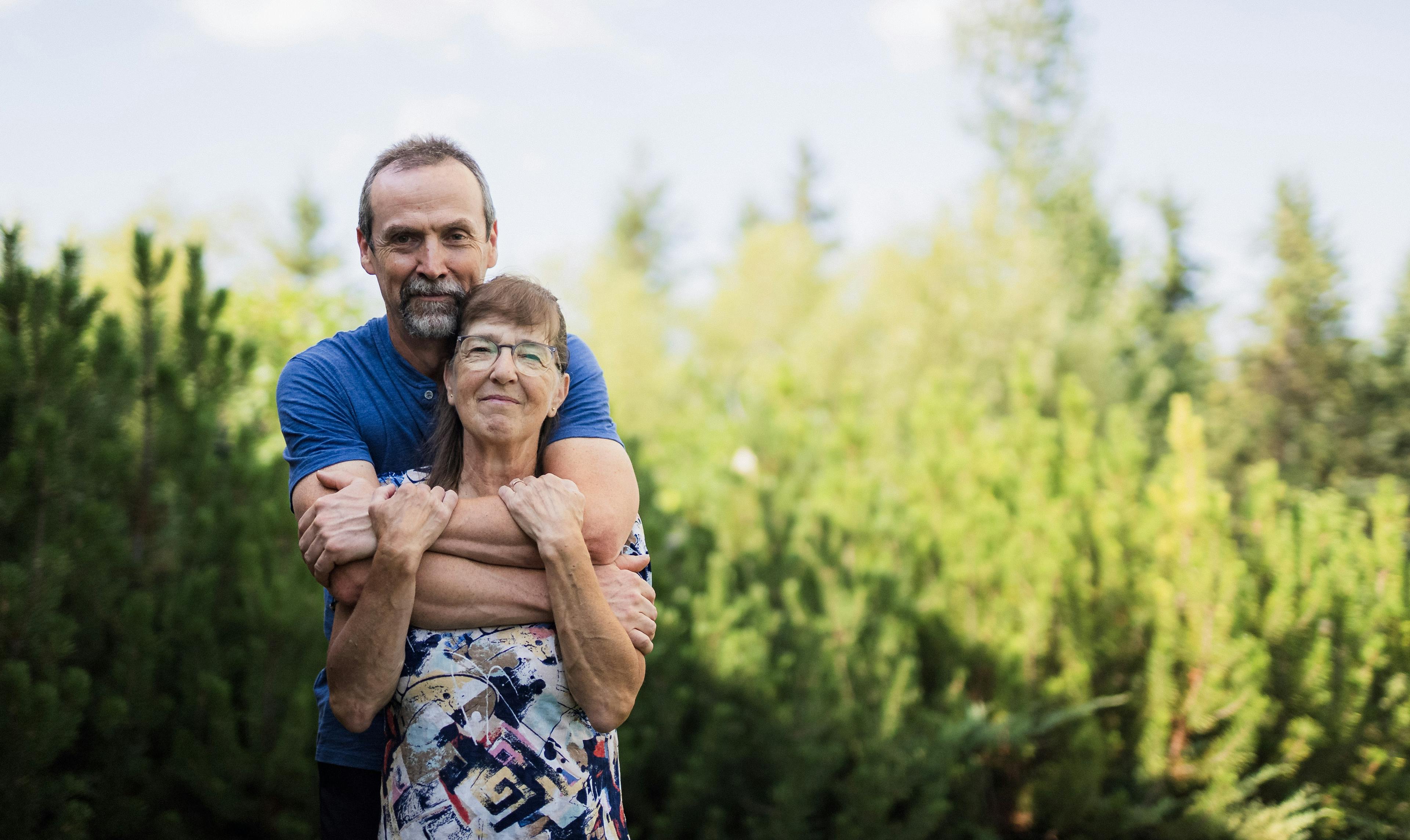 Lorna, who lives with CF, poses with her husband outside of their home in Saskatchewan, surrounded by trees.