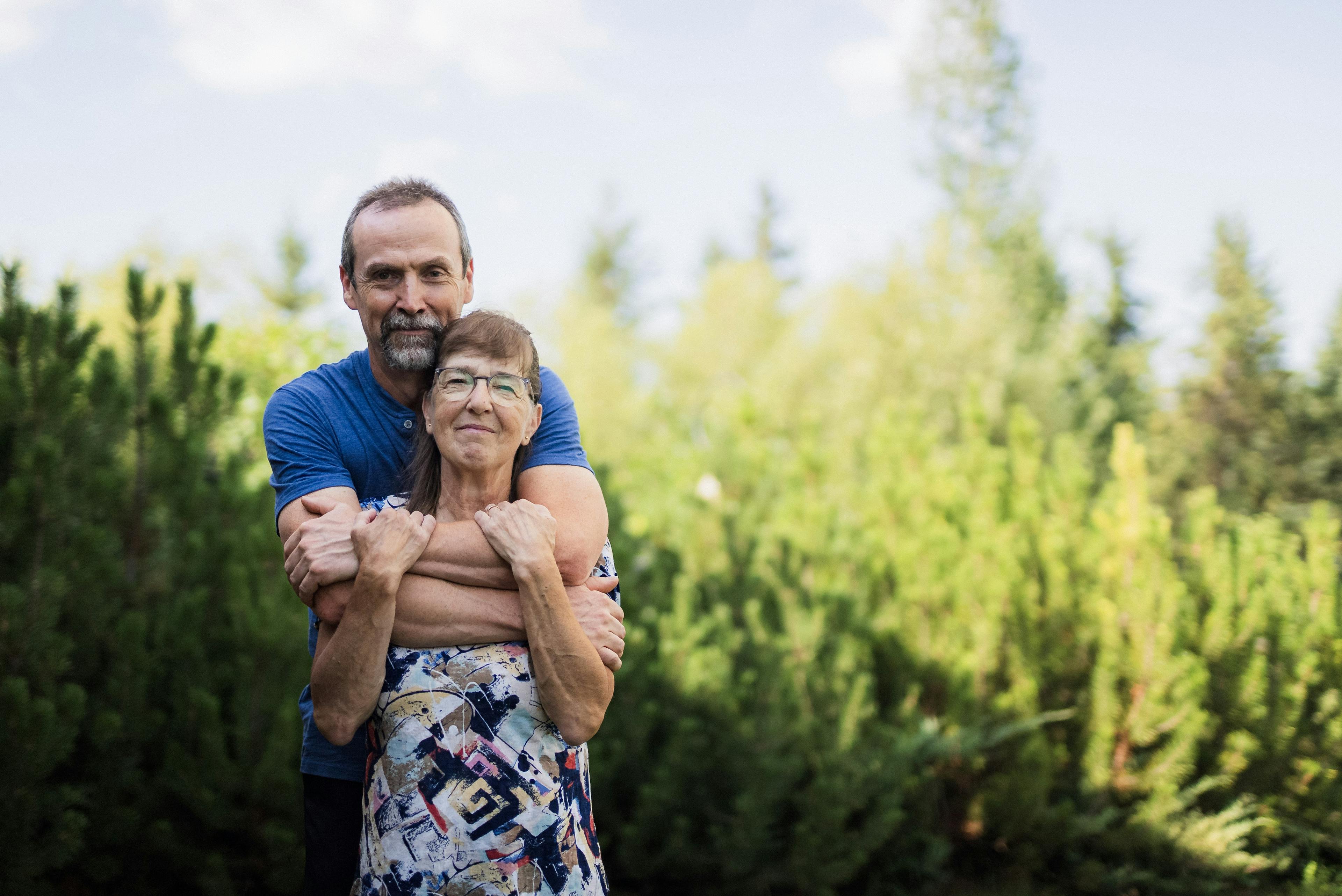 Lorna, who lives with CF, poses with her husband outside of their home in Saskatchewan, surrounded by trees. 