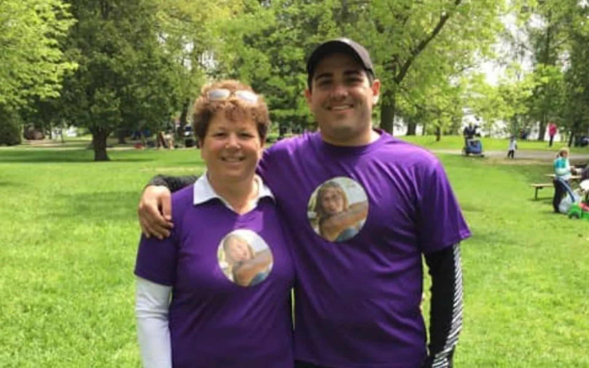 A mother and son posing in a park, wearing purple t-shirts with a young woman's face on them.