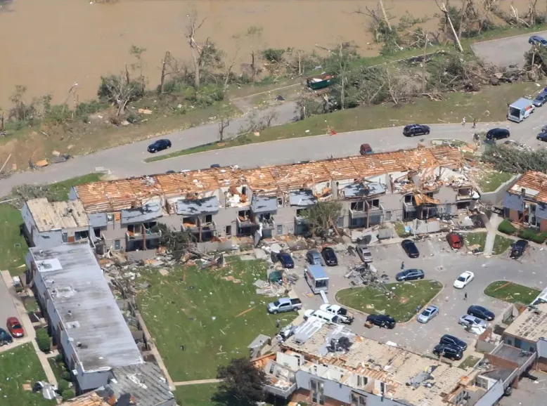 Aerial view of a Dayton apartment complex with its roofs torn off and debris scattered across the grounds after the May 27, 2019 tornado, with a flooded river behind it. Caption: An apartment complex in Dayton with its roofs torn off after the May 27, 2019 tornado. Many such units were never rebuilt as affordable housing.