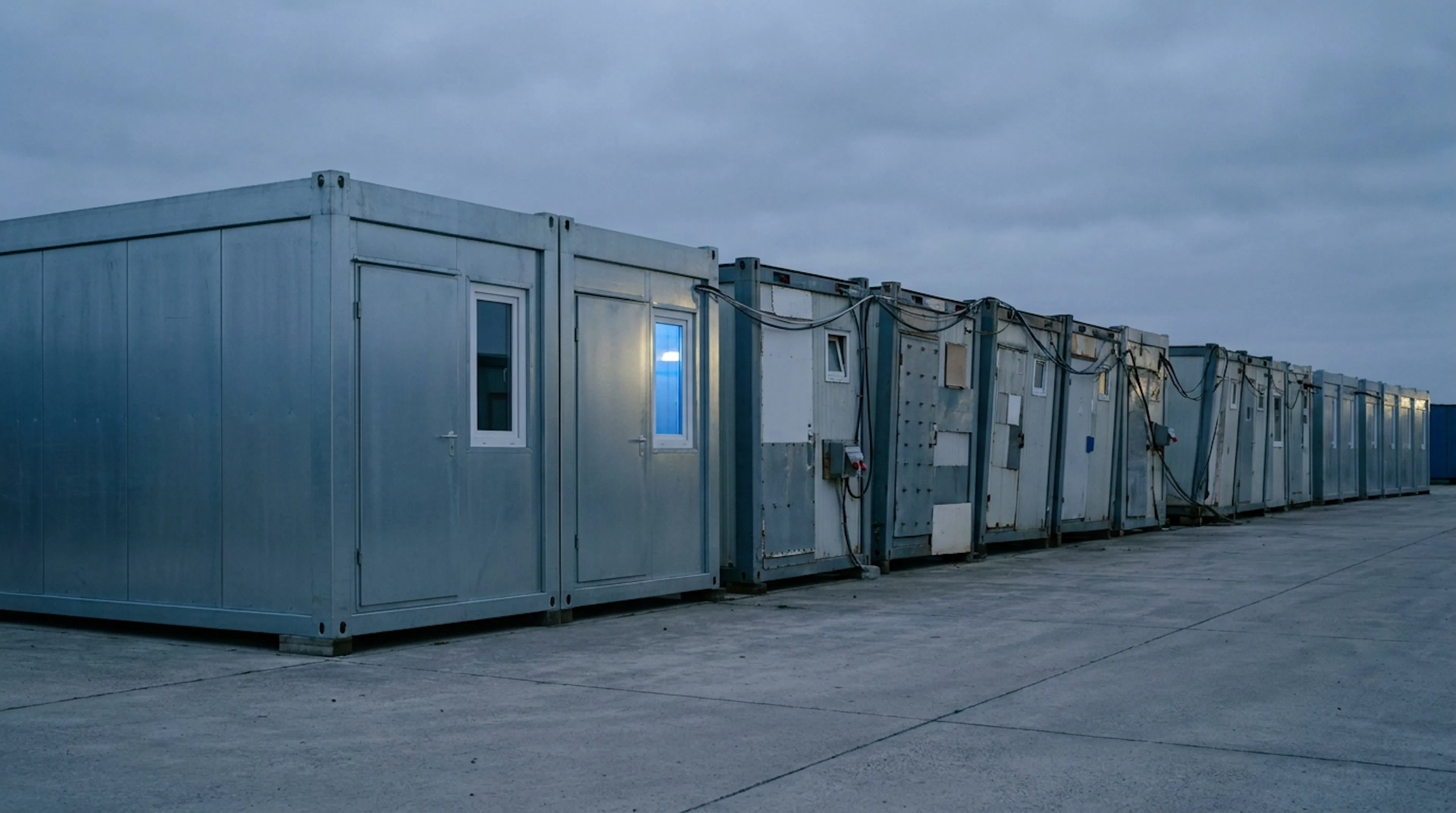 A row of identical prefabricated modular units, pristine at the near end and visibly improvised and strained further down the row